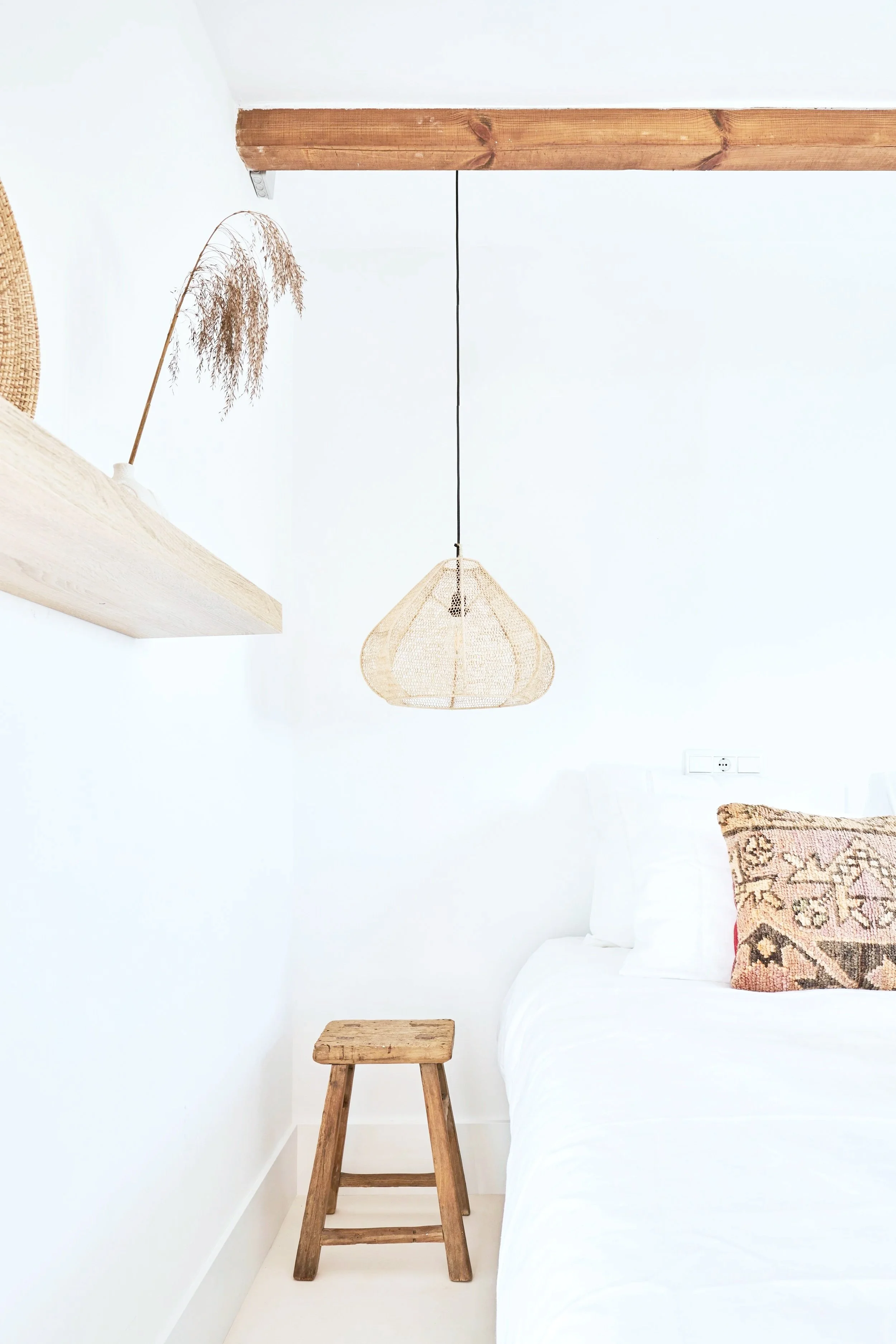 A minimalist bedroom corner with a white wall, a wooden stool, a bed with white bedding and decorative pillows, a hanging woven light fixture, a wall socket, and a wooden beam on the ceiling.
