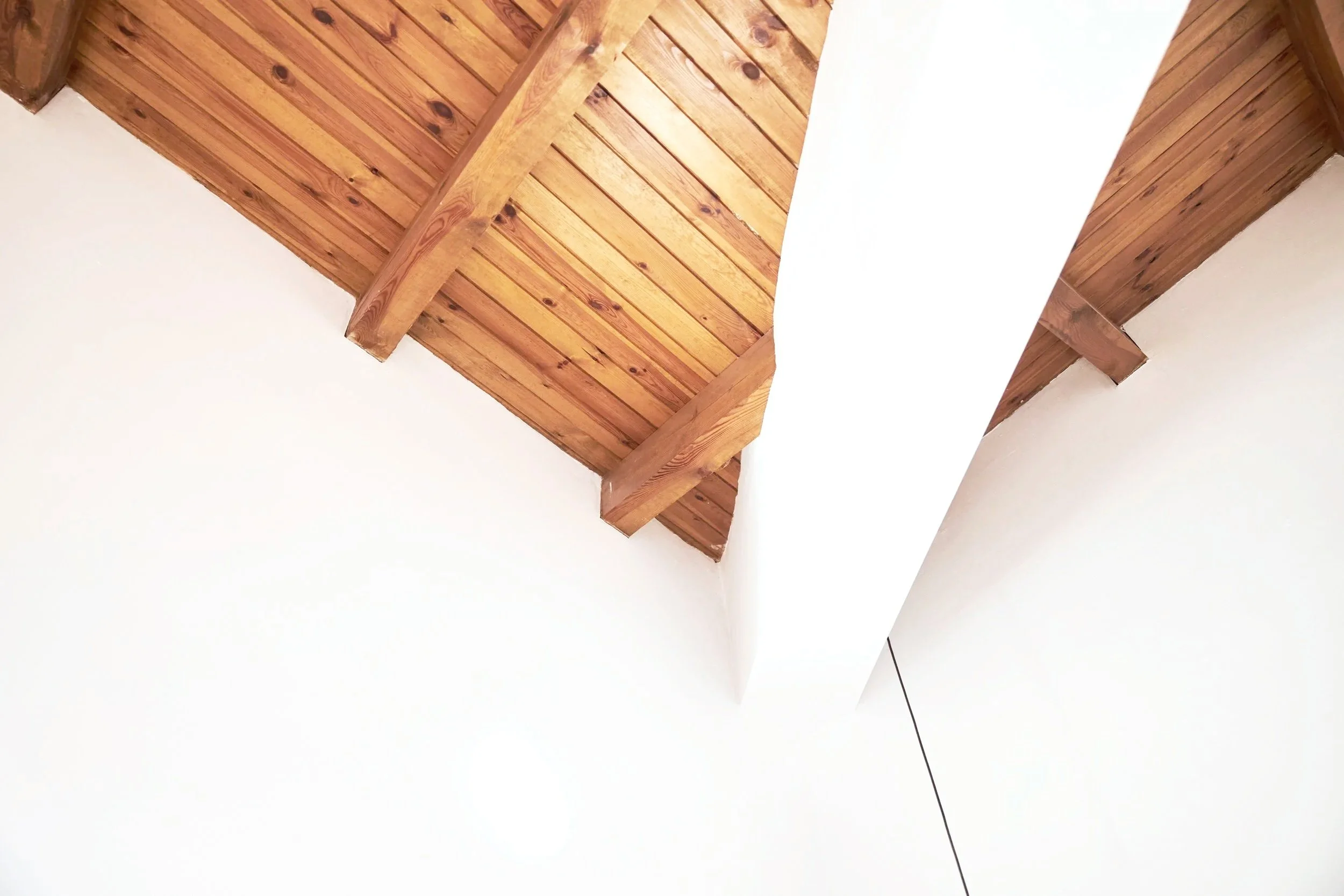 Ceiling with wooden beams and panels, white wall with a circular light fixture, and a white crossbeam.