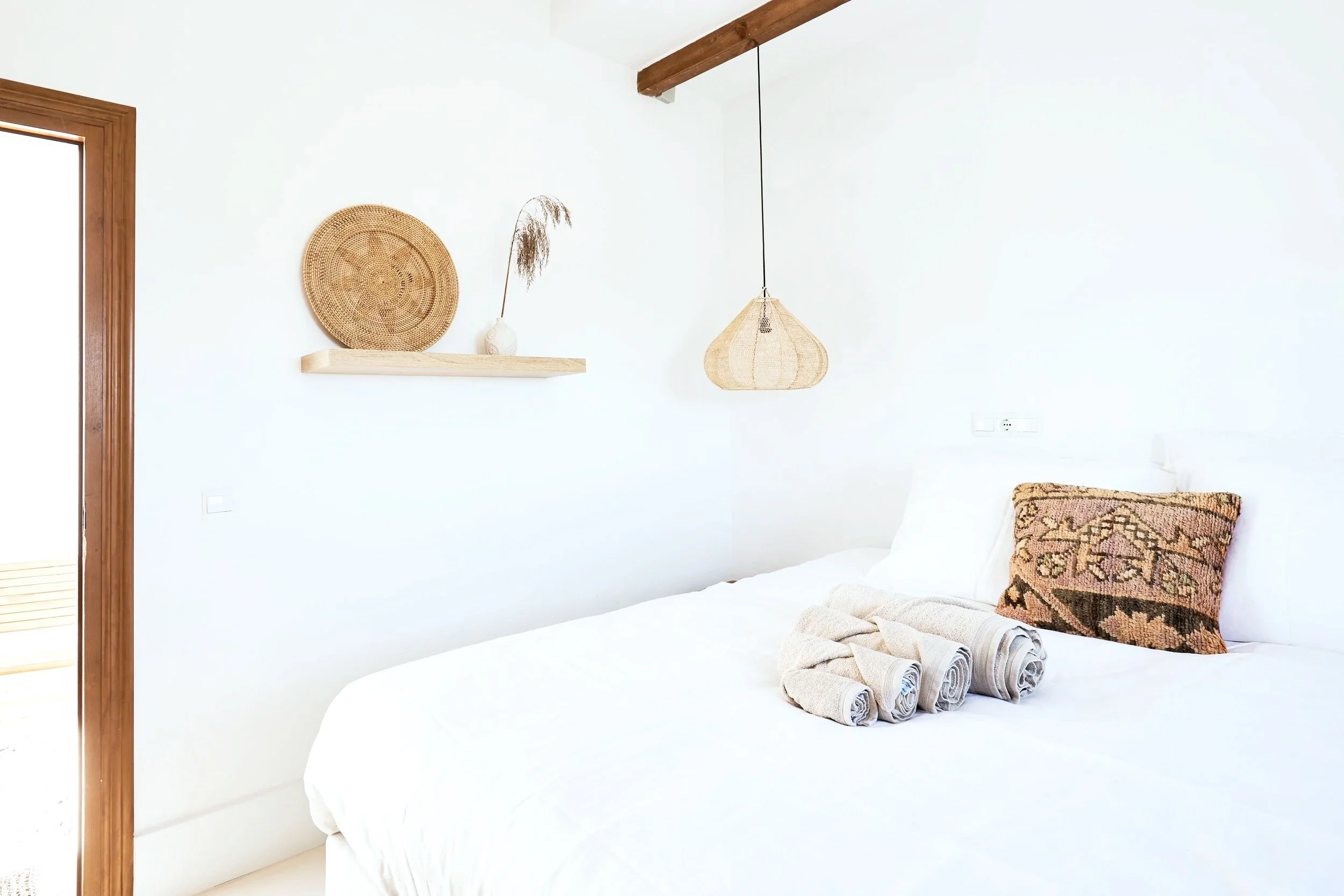A neatly made bed with white linens, inching pillows, and decorative rolled towels, with a rustic woven pillow, against a white wall. On the wall, there is a floating shelf with decorative baskets and a vase with dried ornamental grass. A hanging wic