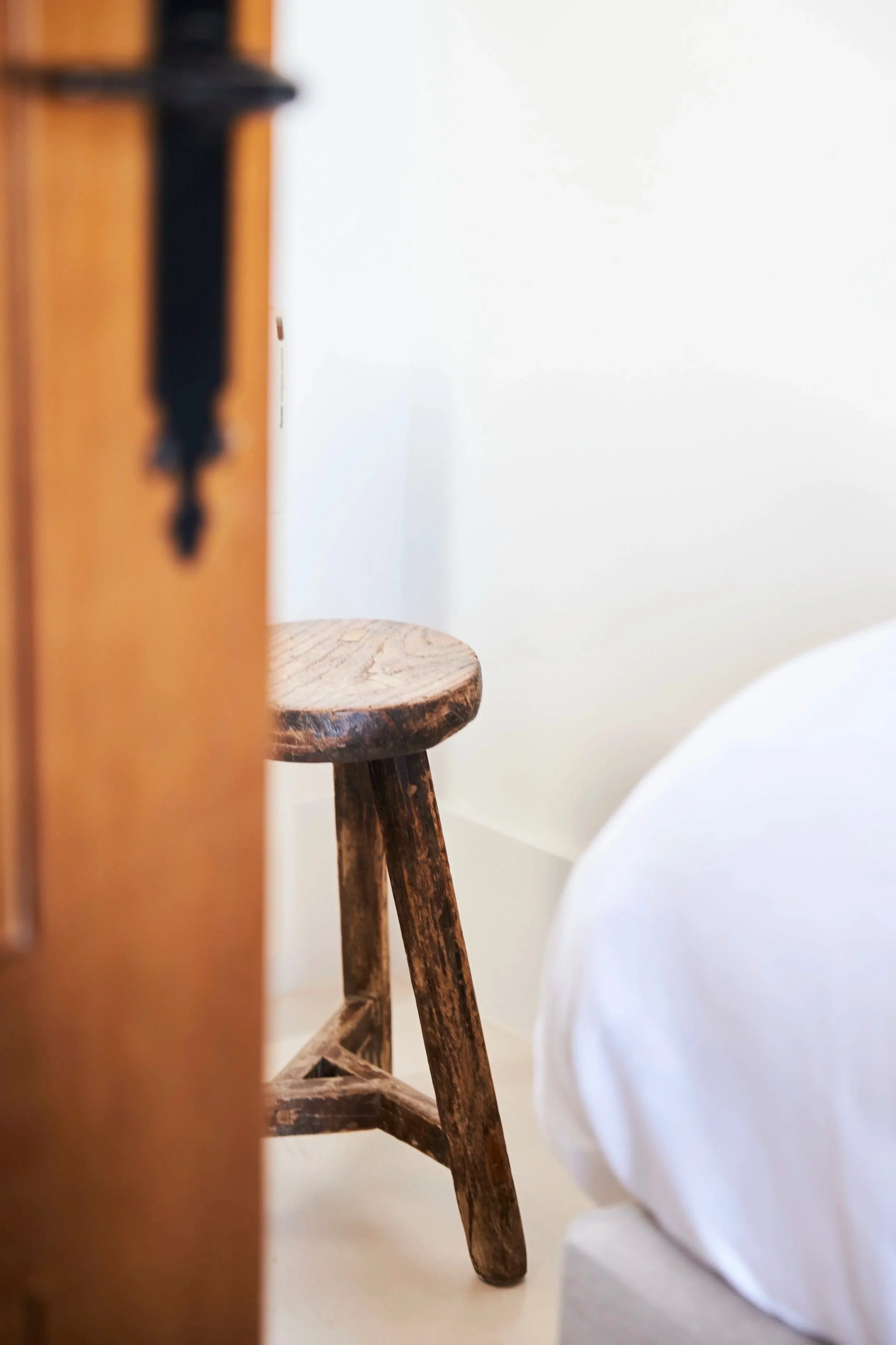 A rustic wooden stool with three legs visible, placed next to a white bed and partially obscured by a blurred wooden piece in the foreground, against a white wall background.
