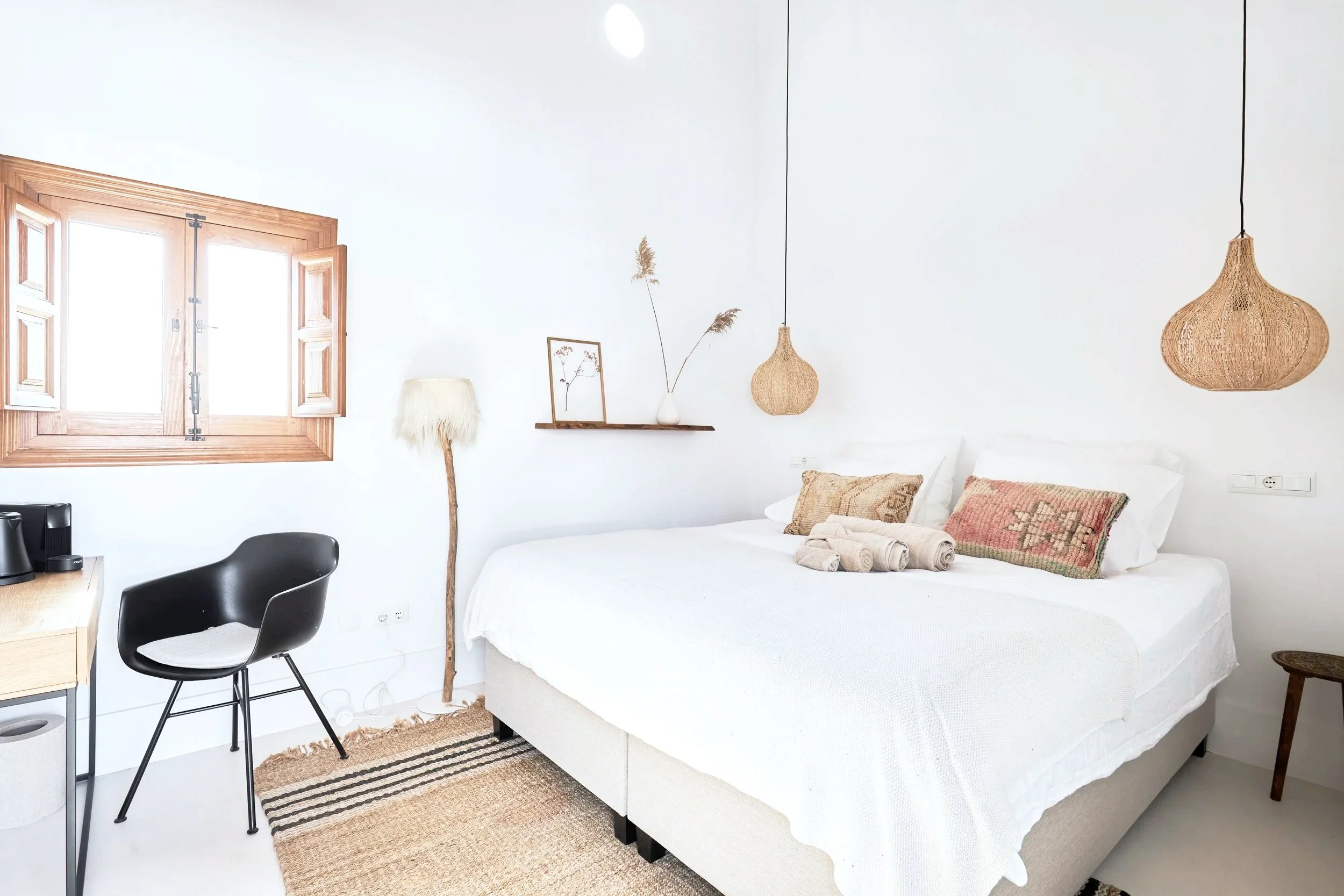 Bright bedroom with a large bed, white bedding, decorative pillows, rolled towels, a wooden side table, a wall-mounted wooden shelf with a vase and dried plants, hanging wicker light fixtures, a window with wooden shutters, a dark chair, a lamp with 
