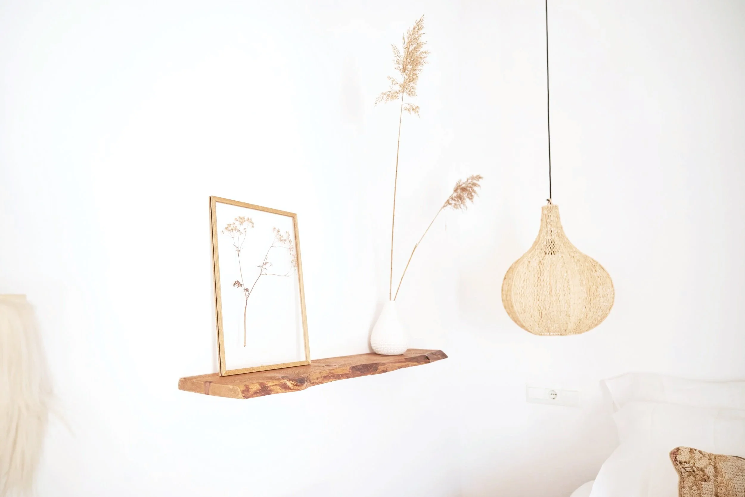 Decorative interior with a white wall, a wooden shelf holding an art piece of a pressed plant in a wooden frame, and a white vase with dried grass. A woven lampshade hangs nearby, and part of a bed with white bedding is visible.
