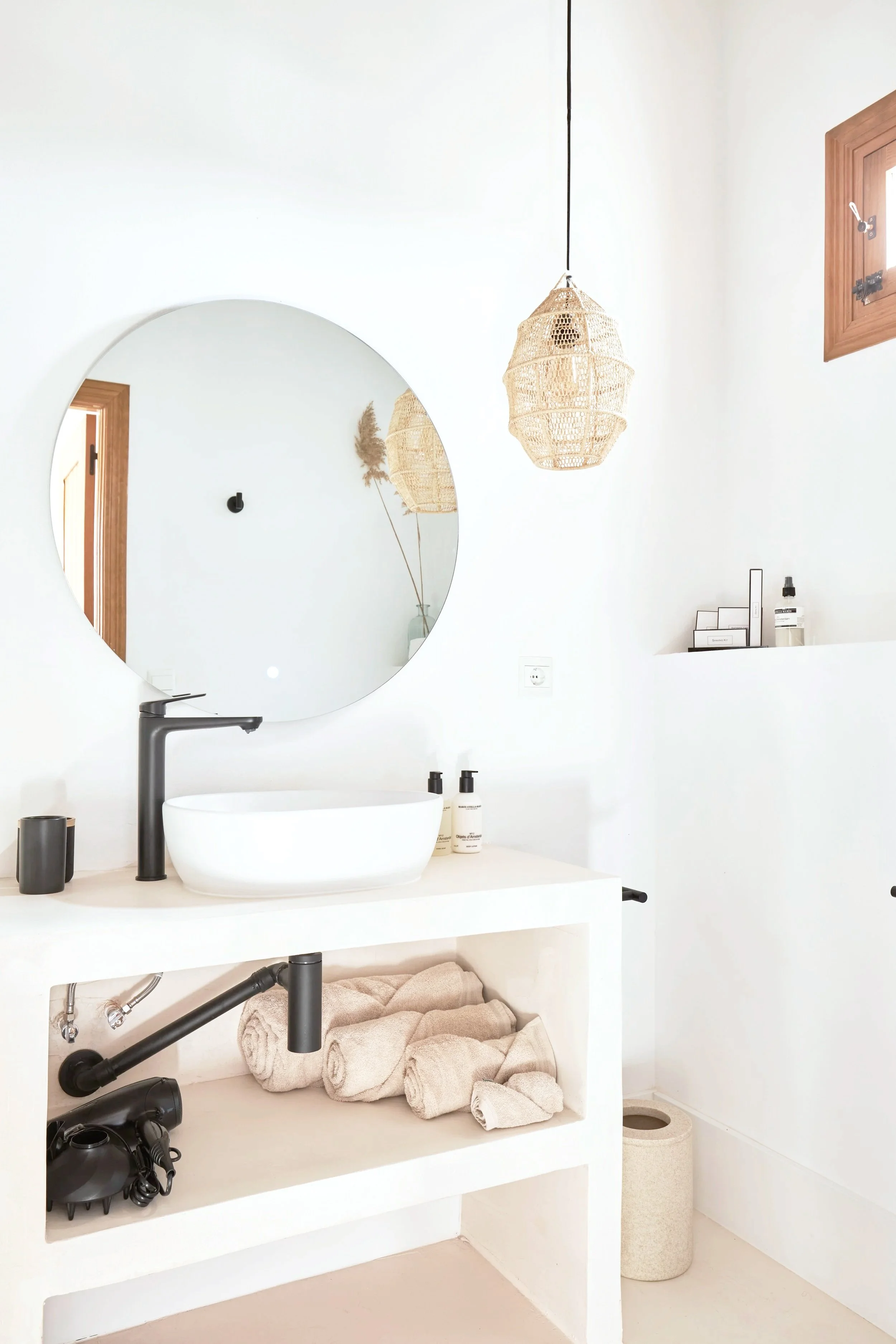 Modern bathroom with a round mirror, black faucet, white vessel sink, beige towels, black hairdryer, and natural woven pendant lights.