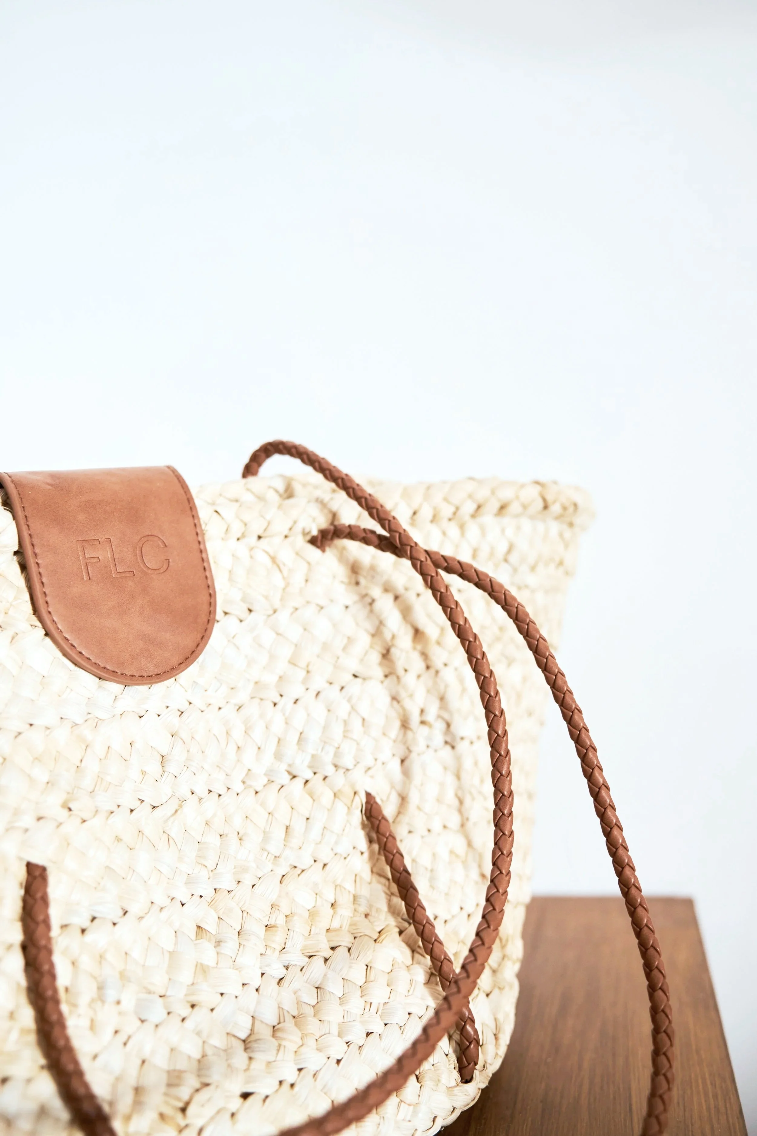 Close-up of a woven straw tote bag with leather accents and braided leather handles on a wooden surface against a plain white background.