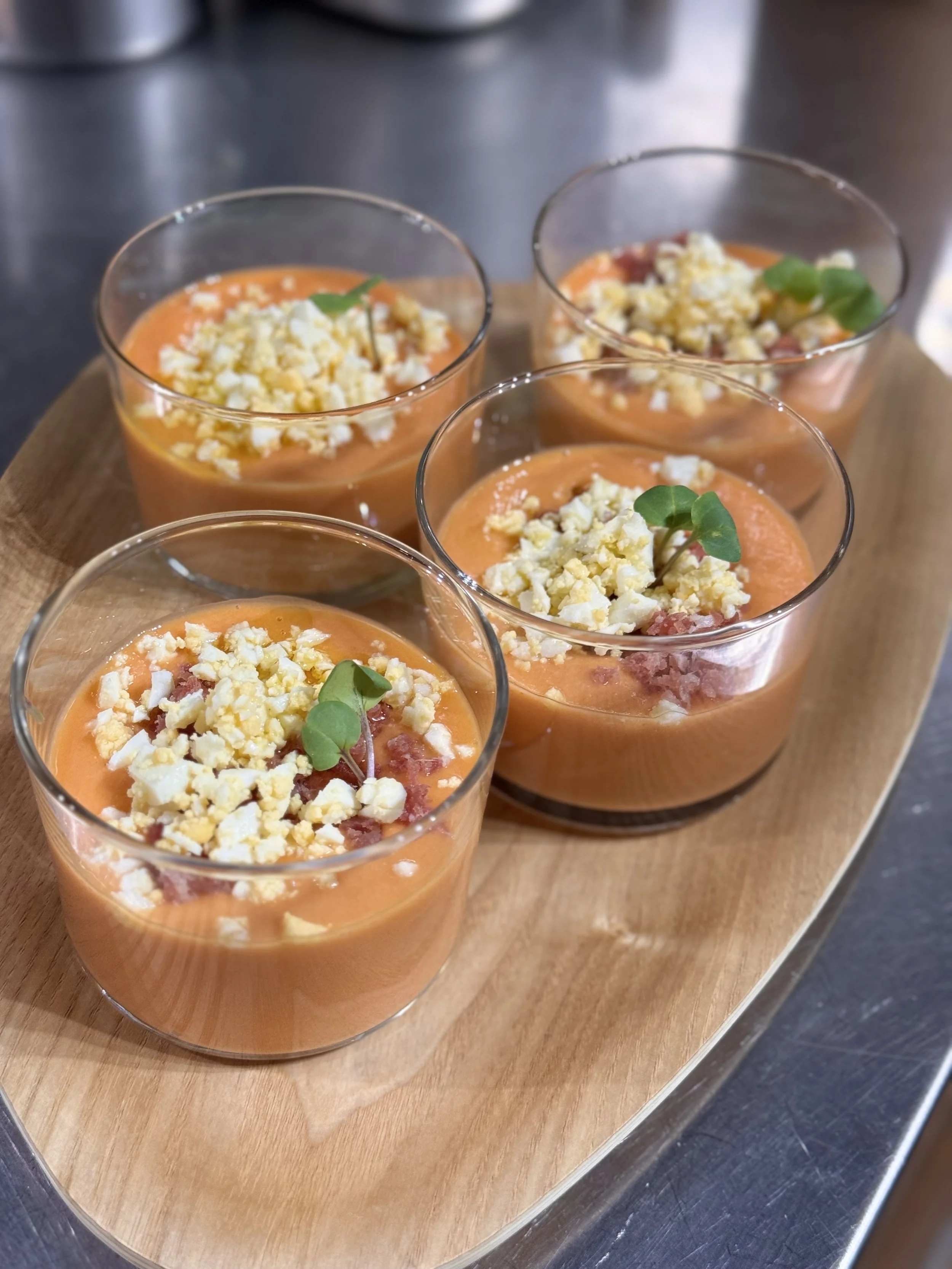 Four glasses of orange-colored soup topped with crumbled cheese and garnished with small herbs, served on a wooden platter.