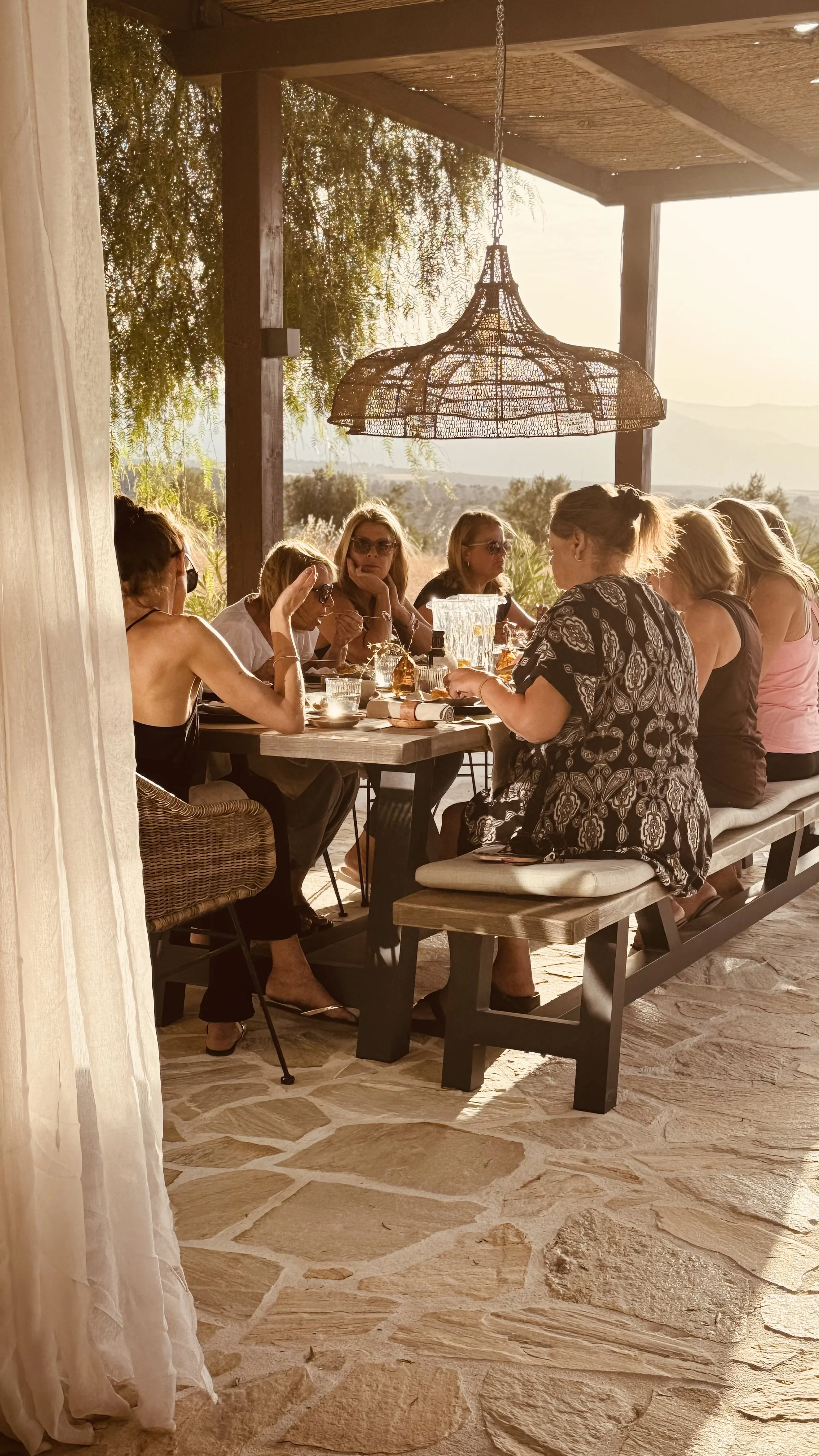 A group of women having a meal outdoors at a rustic dining area with a chandelier, surrounded by nature during sunset.
