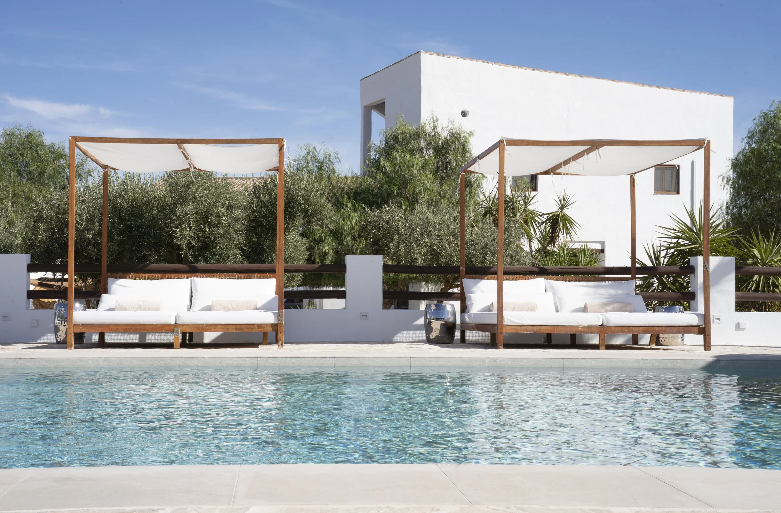 Two white canopy lounge beds with white cushions by a swimming pool, with greenery and a white building in the background under a clear blue sky.