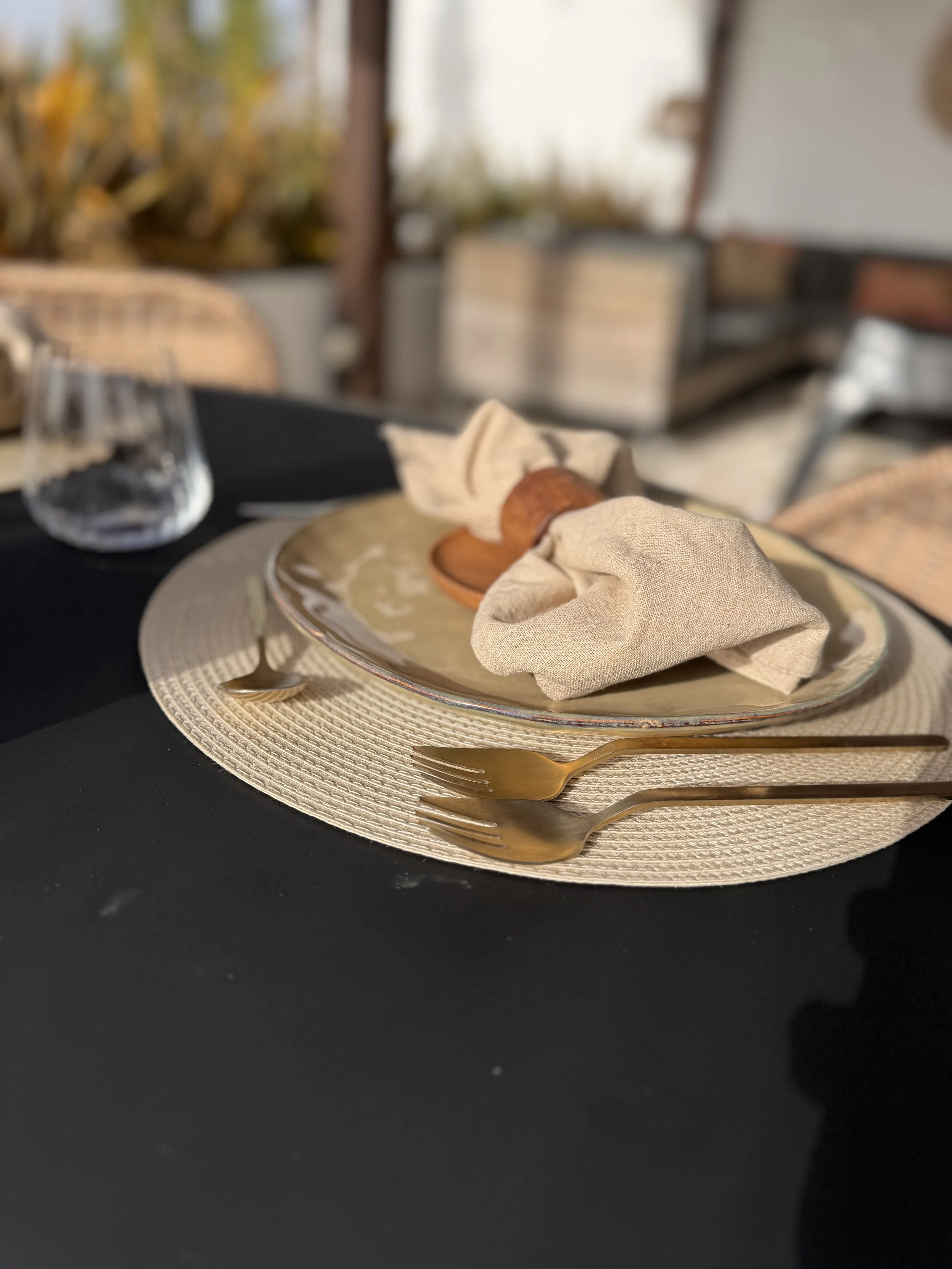A dining table set with a round woven placemat, gold-colored forks, a beige cloth napkin, a plate with a bread roll, and a glass of water, with blurred outdoor background.