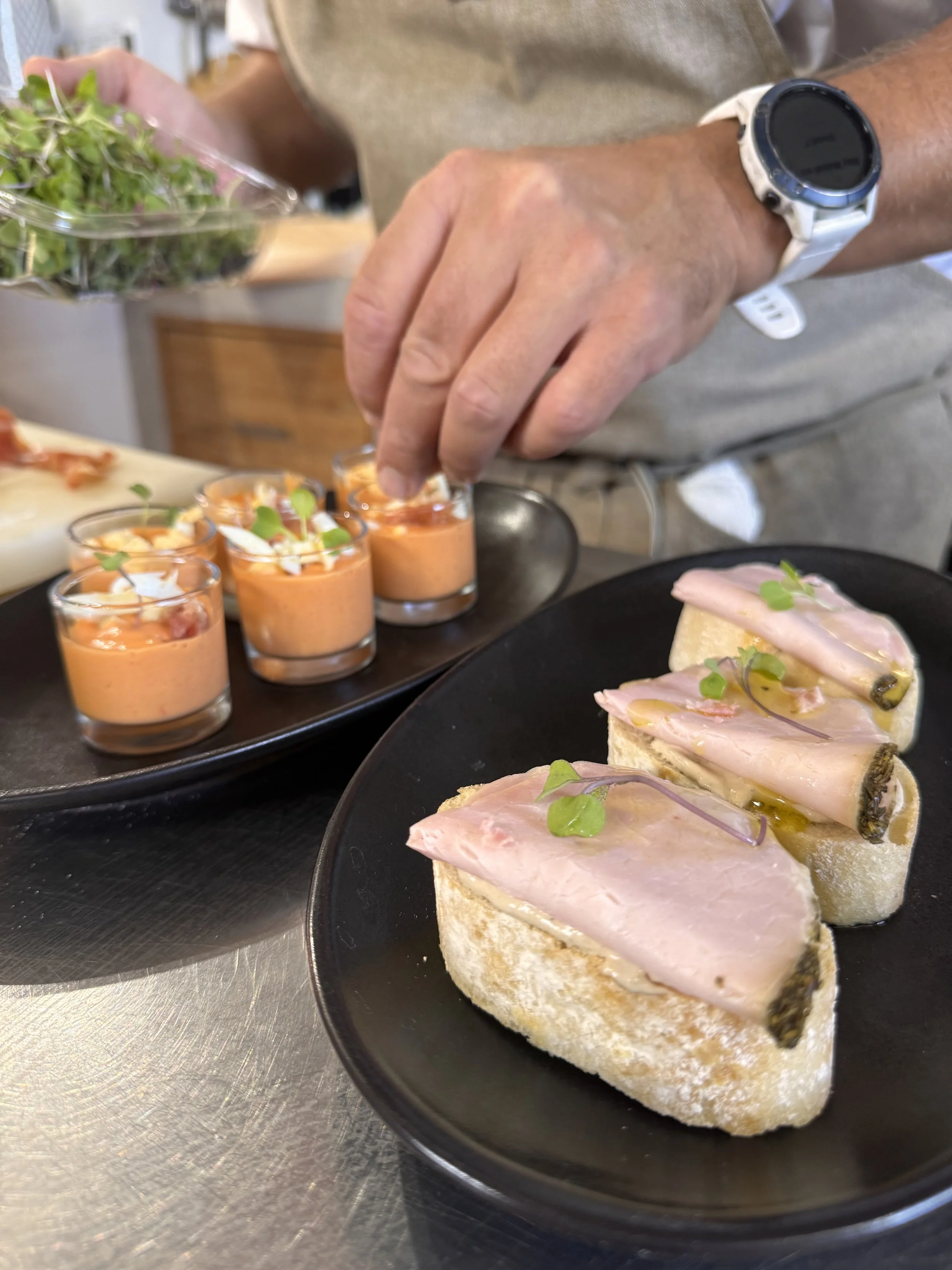 Close-up of a chef garnishing small open-faced sandwiches with microgreens and a row of shrimp toast with microgreens on a black plate, with small cups of shrimp cocktail in the background.