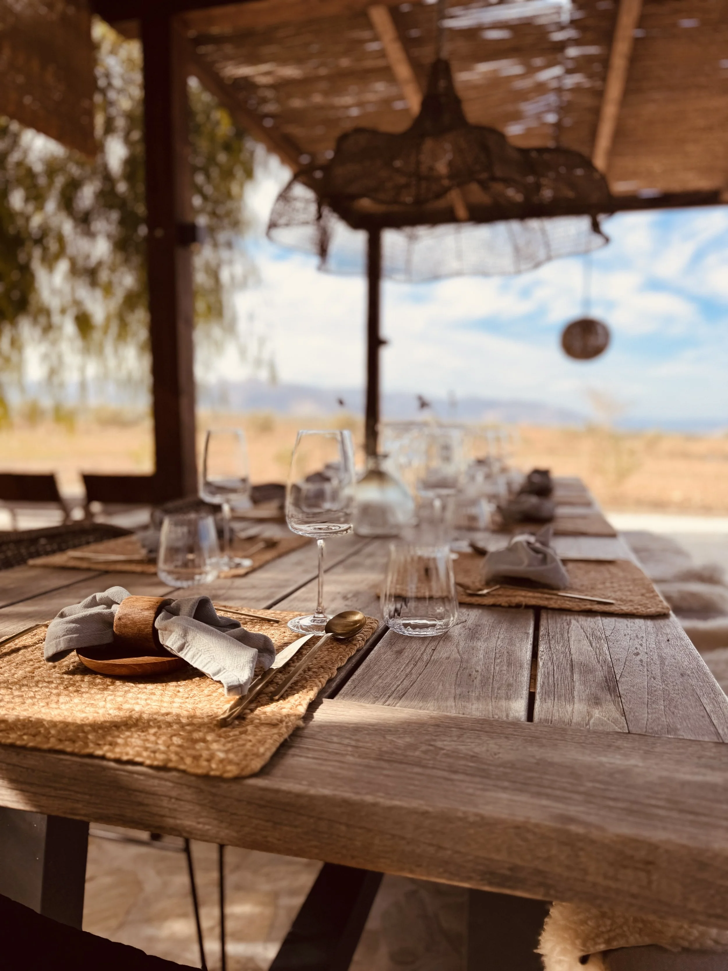 Outdoor dining table set with wine glasses, water glasses, woven placemats, napkins, and cutlery, under a wooden roof with a seaside landscape in the background.