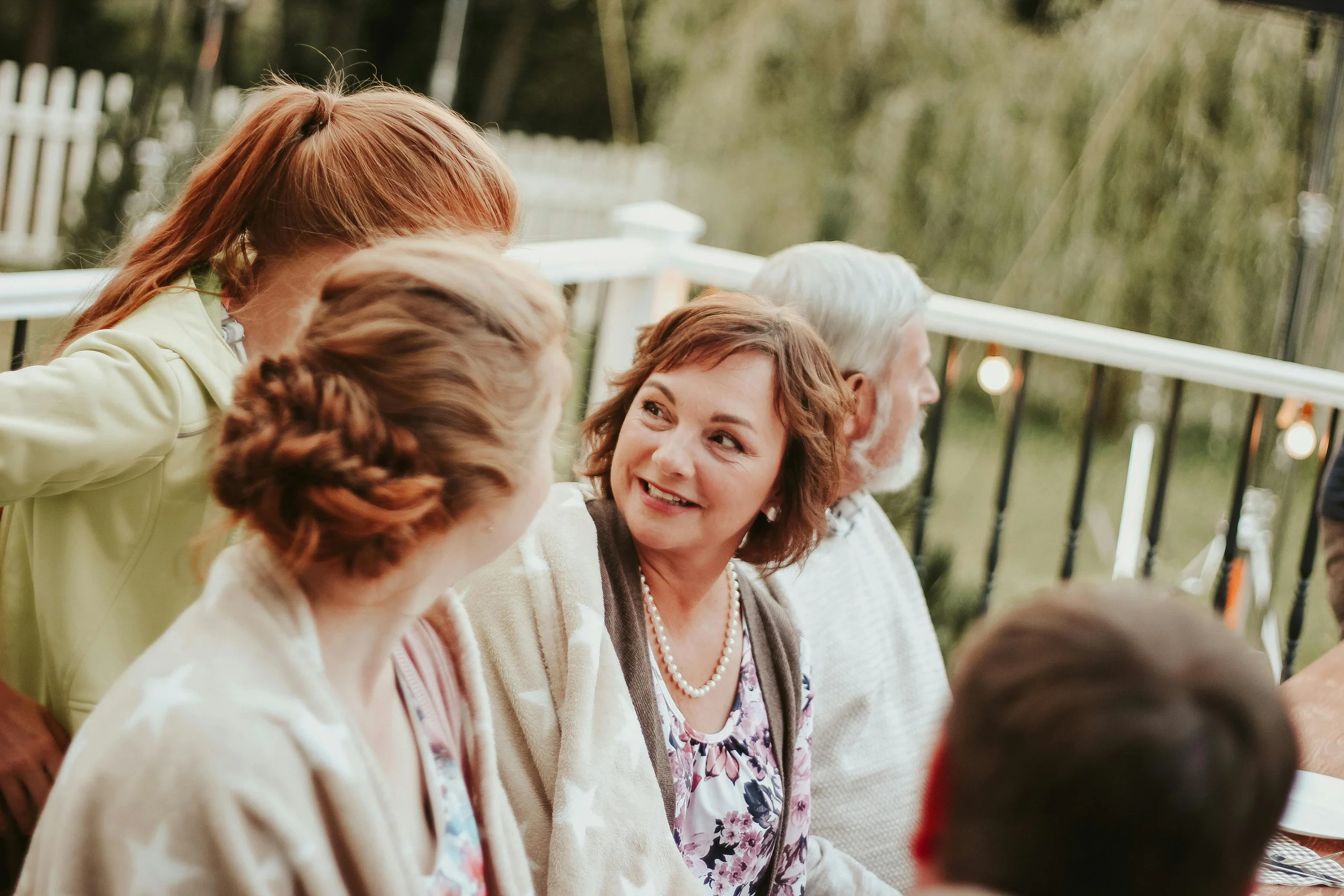 A group of people sitting and talking outdoors on a patio, with a woman in the center smiling and engaging in conversation.