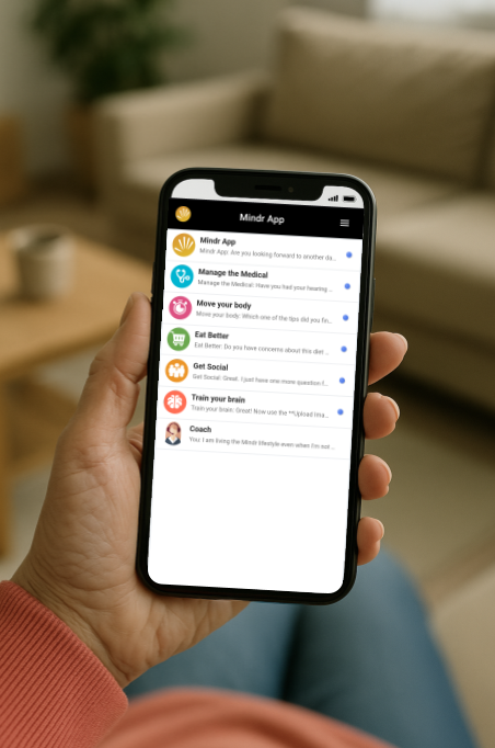 Hand holding smartphone displaying a wellness app with options like Mindr App, Manage the Medical, Move your body, Eat Better, Get Social, Train your brain, and Coach, in a cozy living room setting.