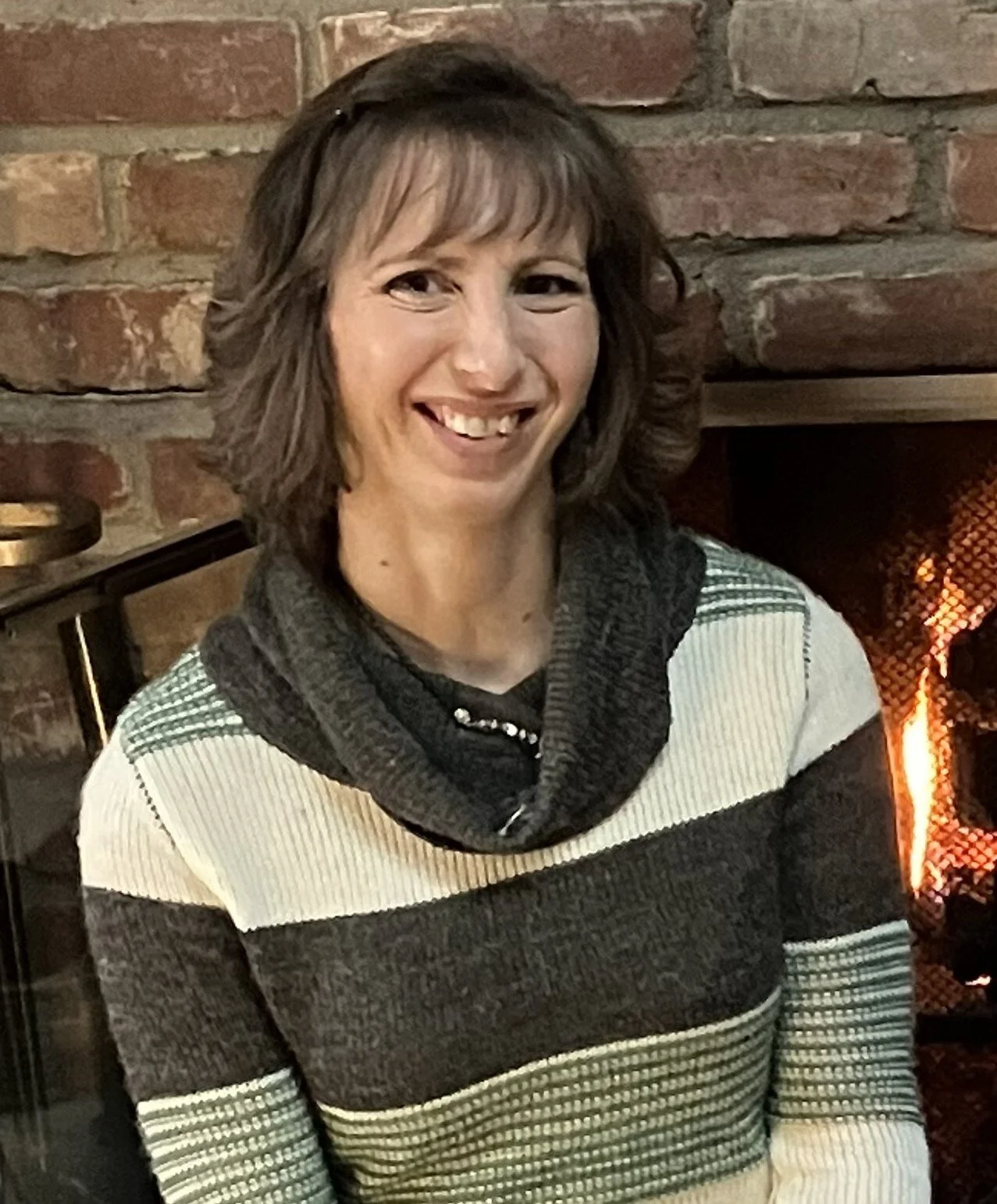 A woman with shoulder-length brown hair and a striped sweater sitting near a brick fireplace.