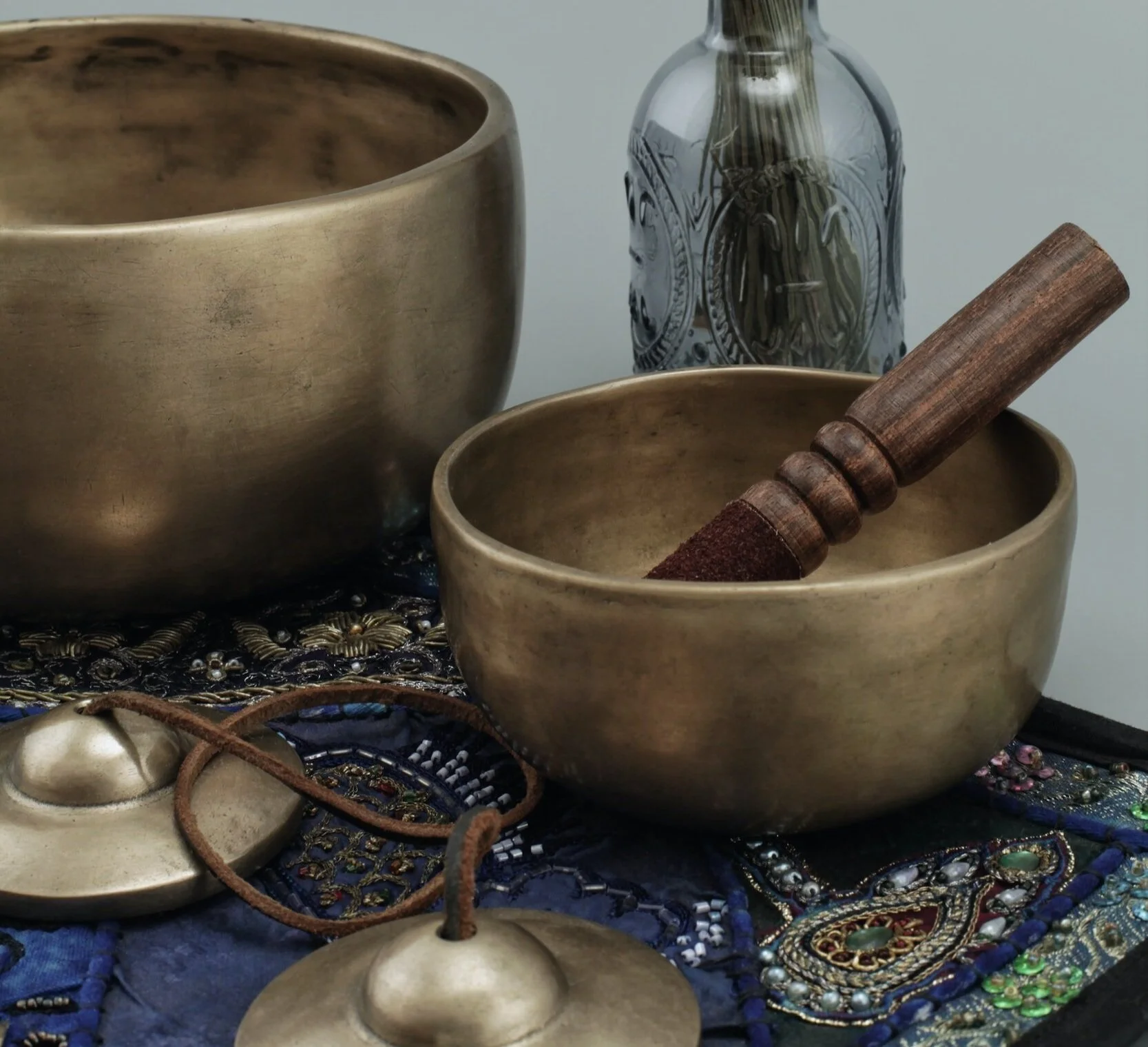 Two brass singing bowls with a wooden mallet inside one bowl, placed on a decorative embroidered fabric with beads and jewelry, with a glass bottle in the background.