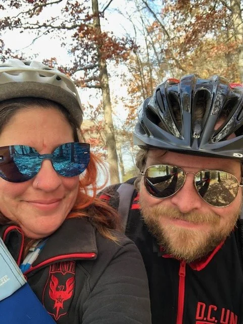 Selfie photo of Marissa and her husband, wearing sunglasses and bike helmets, with Rees behind them.