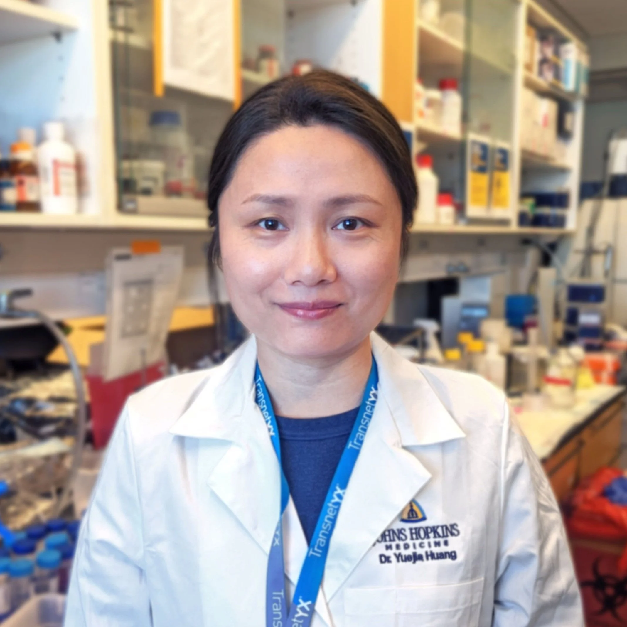 A woman in a white lab coat with a blue lanyard stands in a laboratory surrounded by scientific equipment and supplies.