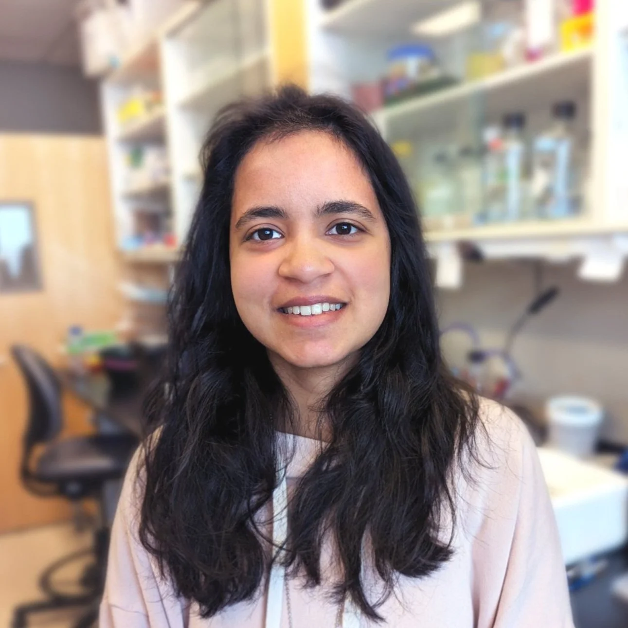 Portrait of a young woman with long dark hair, smiling at the camera, in a laboratory setting.