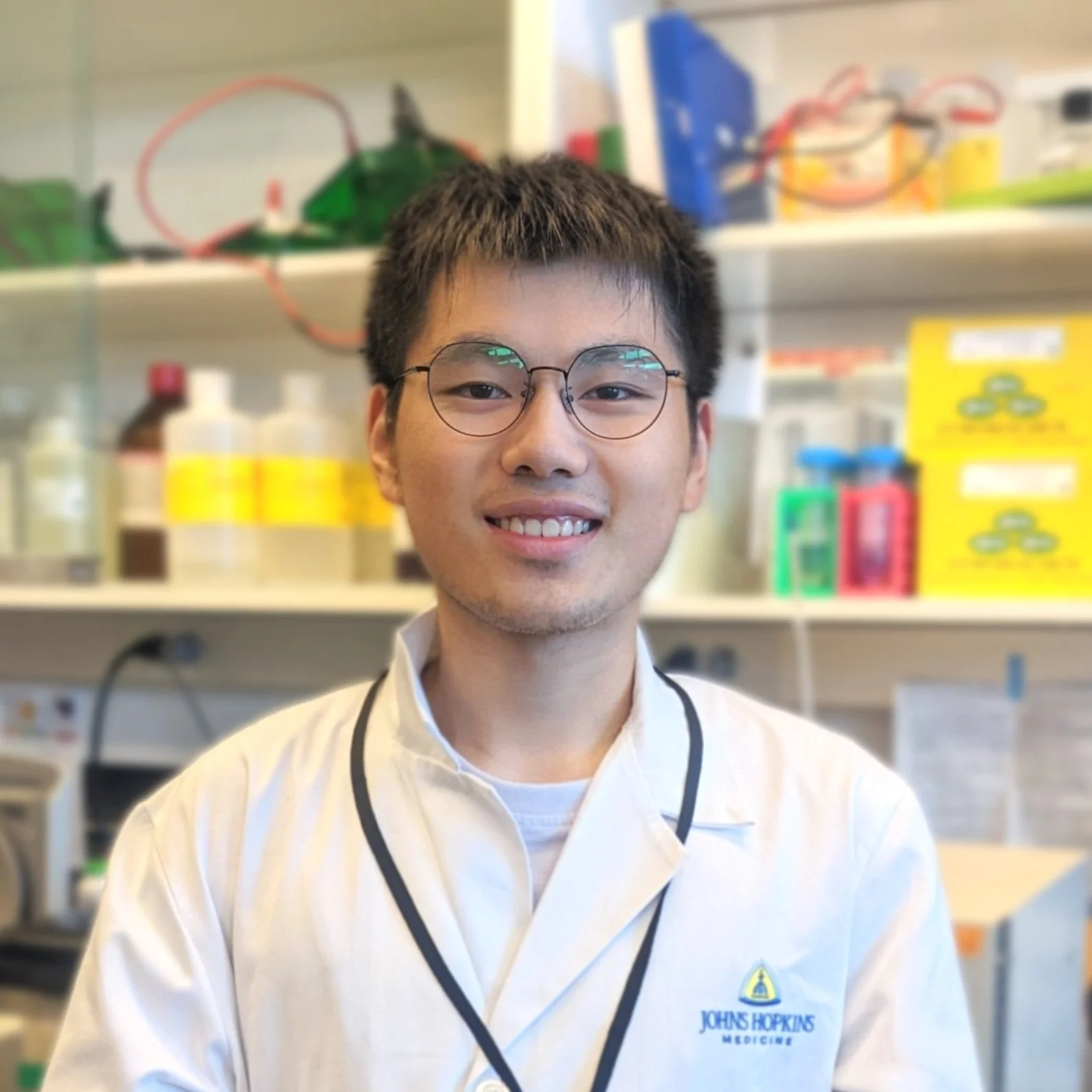 A young man with short dark hair, round glasses, and a cheerful smile, wearing a white lab coat with Johns Hopkins Medicine logo, standing in a laboratory with shelves of lab supplies and chemicals behind him.