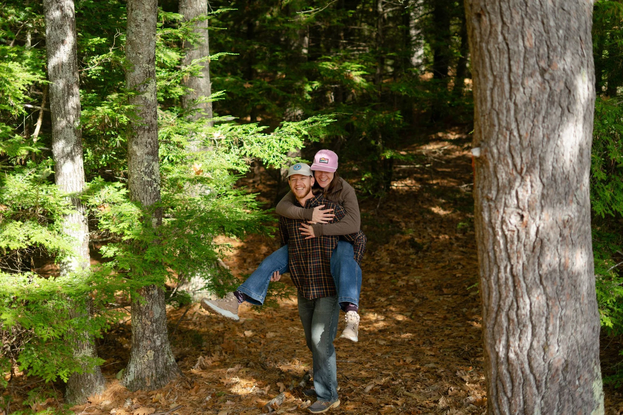 A happy couple in casual clothing and hats enjoying a piggyback ride through a lush green forest.