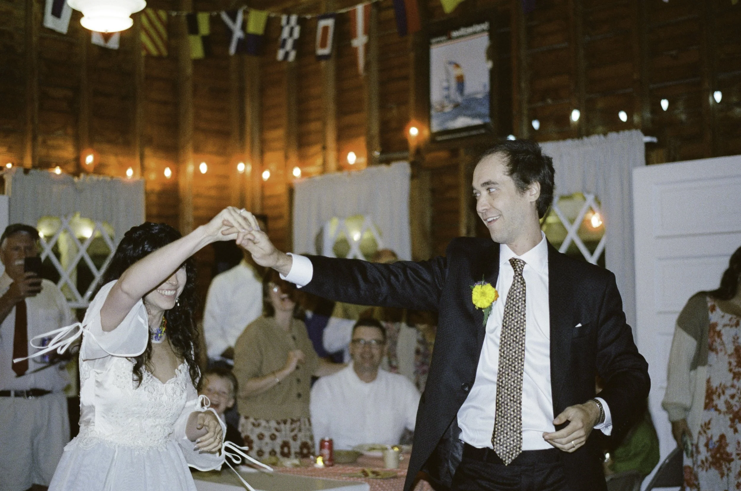 A groom and bride dancing at a wedding reception with guests watching and taking photos in the background. The venue has wooden walls and decorative banners.
