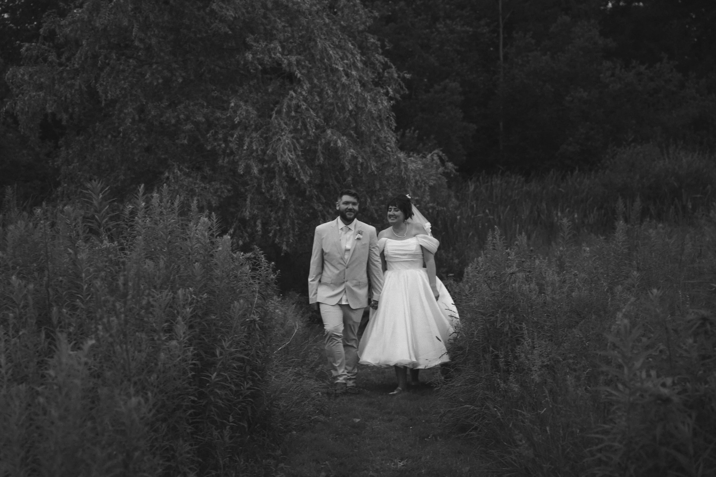 A black and white photograph of a smiling couple, dressed in wedding attire, walking hand in hand through a natural, outdoor setting with tall grass and trees.