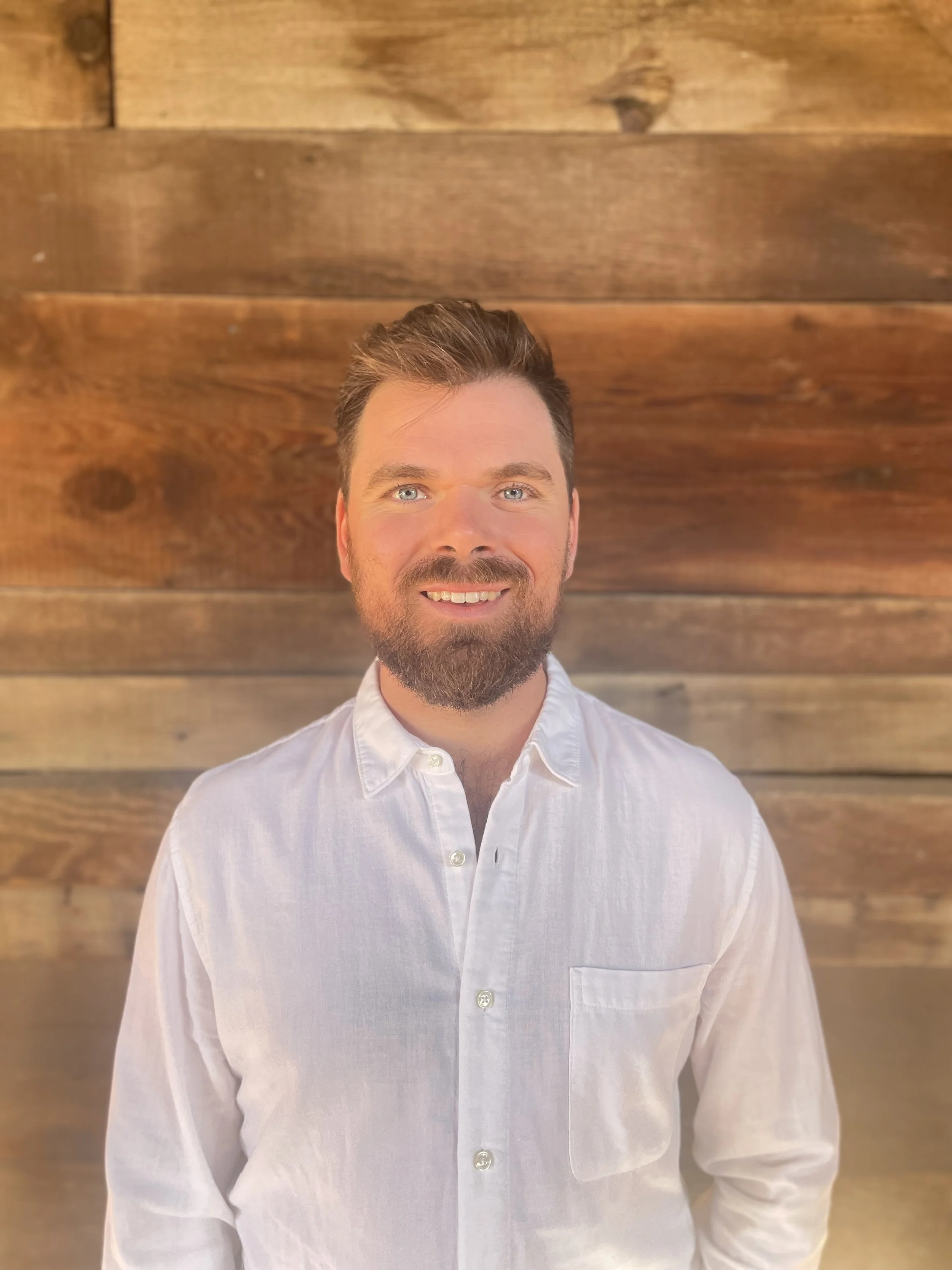 A man with a beard and short brown hair, smiling and wearing a white button-down shirt, standing in front of a wooden wall.