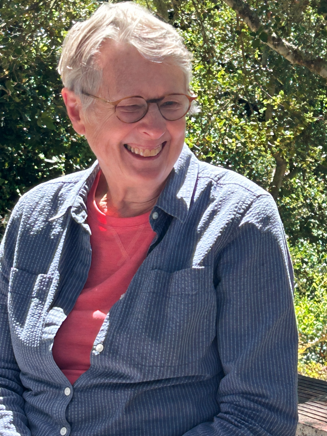 An elderly woman with short gray hair, wearing glasses, a dark striped button-up shirt over a pink t-shirt, smiling and looking to her right outdoors with green foliage and sunlight in the background.