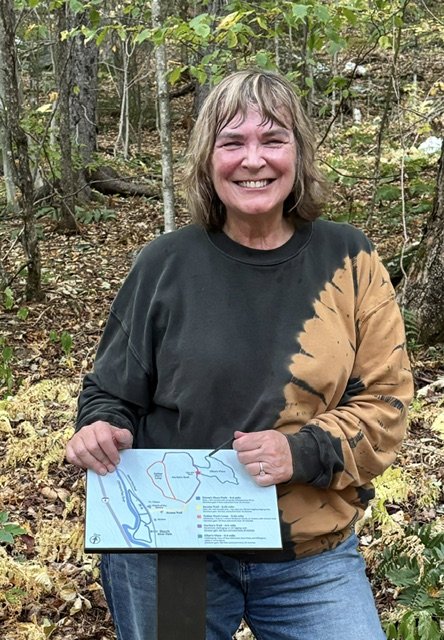 A smiling woman standing in a forest holding a map of a park or nature area with trees and leaves in the background.