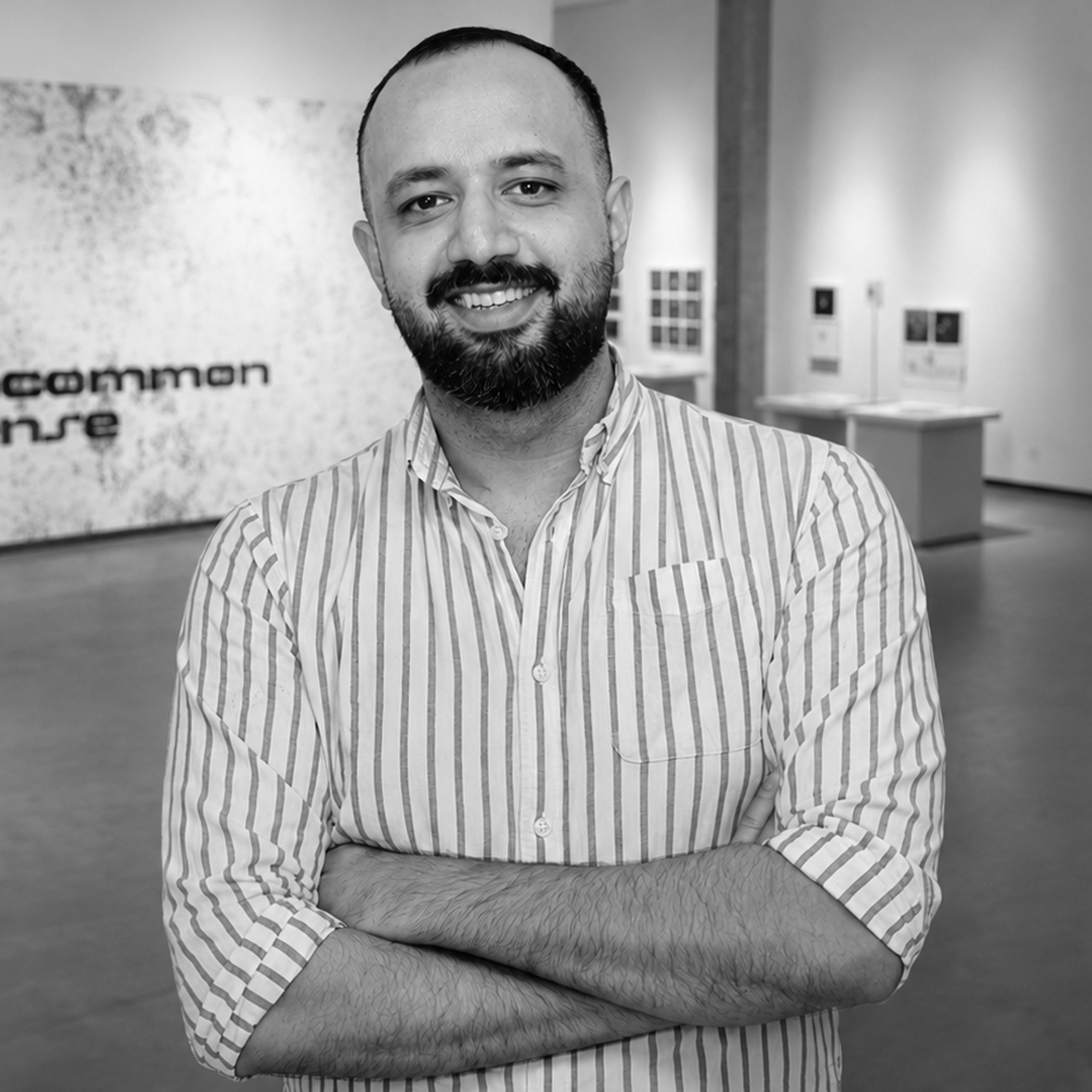 A black and white photo of a man with a beard and mustache, wearing a striped button-up shirt, smiling and posing with arms crossed in an indoor setting with framed pictures or artwork in the background.