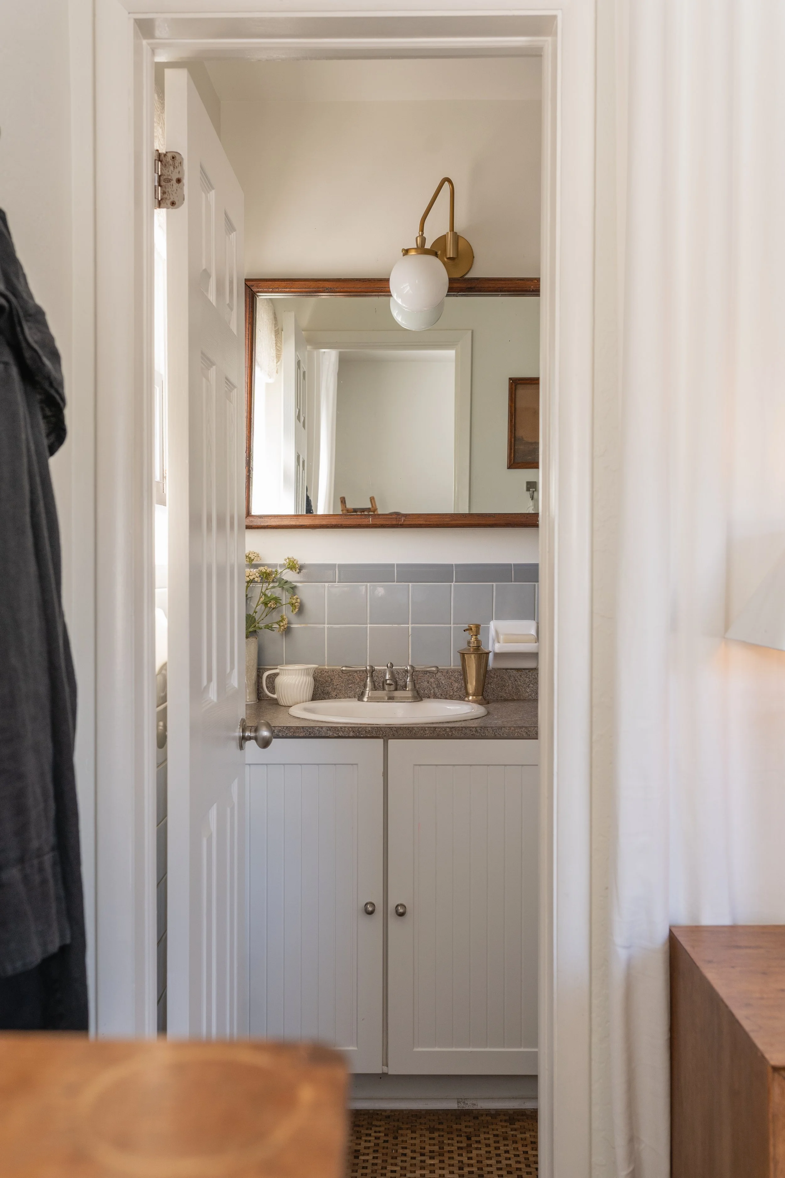 A small bathroom with a white vanity, grey tiled backsplash, and a mirror. A light fixture with a frosted globe is mounted above the mirror. Decorative items and a plant sit on the countertop.