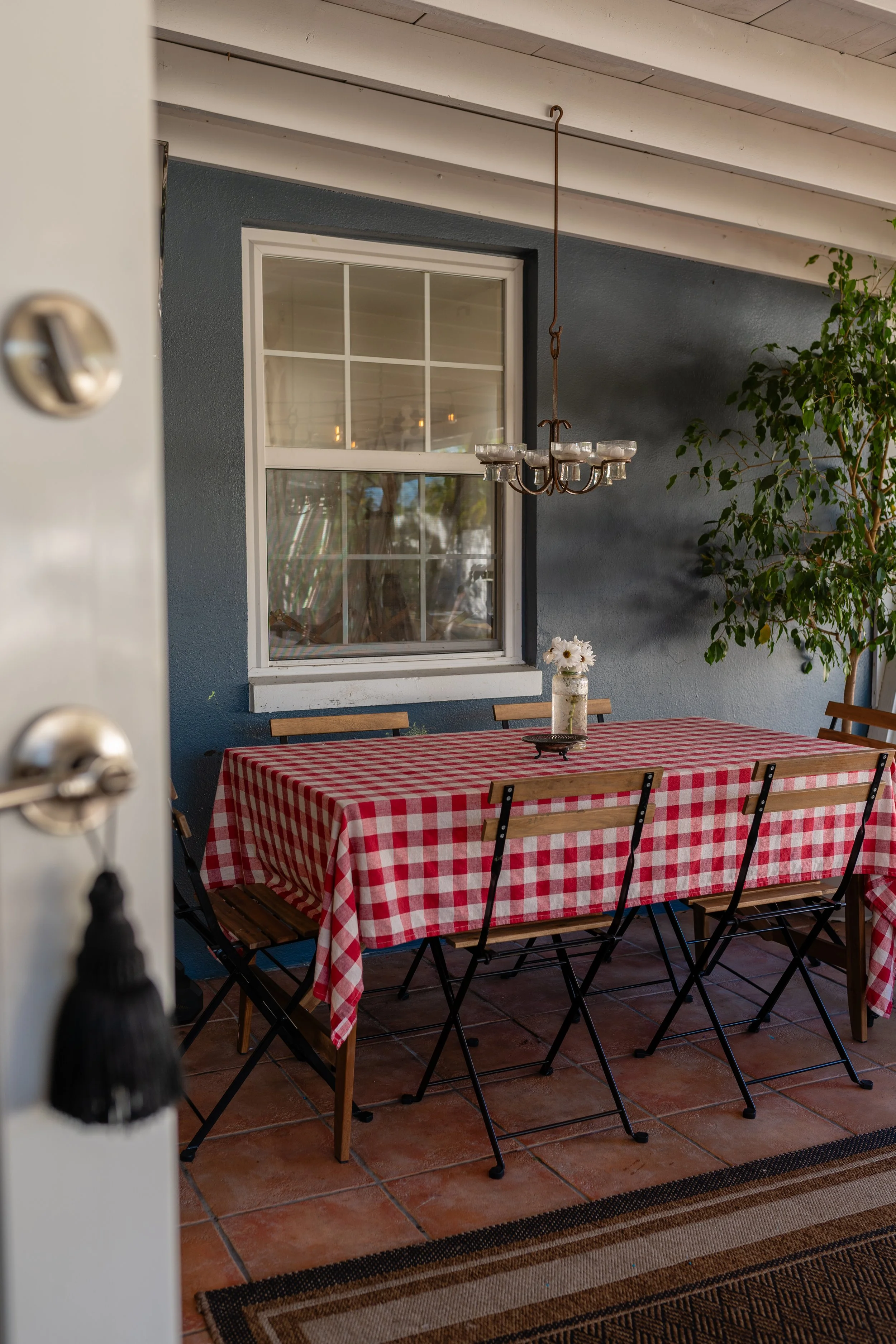 Dining area with a red and white checkered tablecloth, wooden chairs, a glass vase with white flowers, decorative chandelier, blue wall, window, and potted plant.
