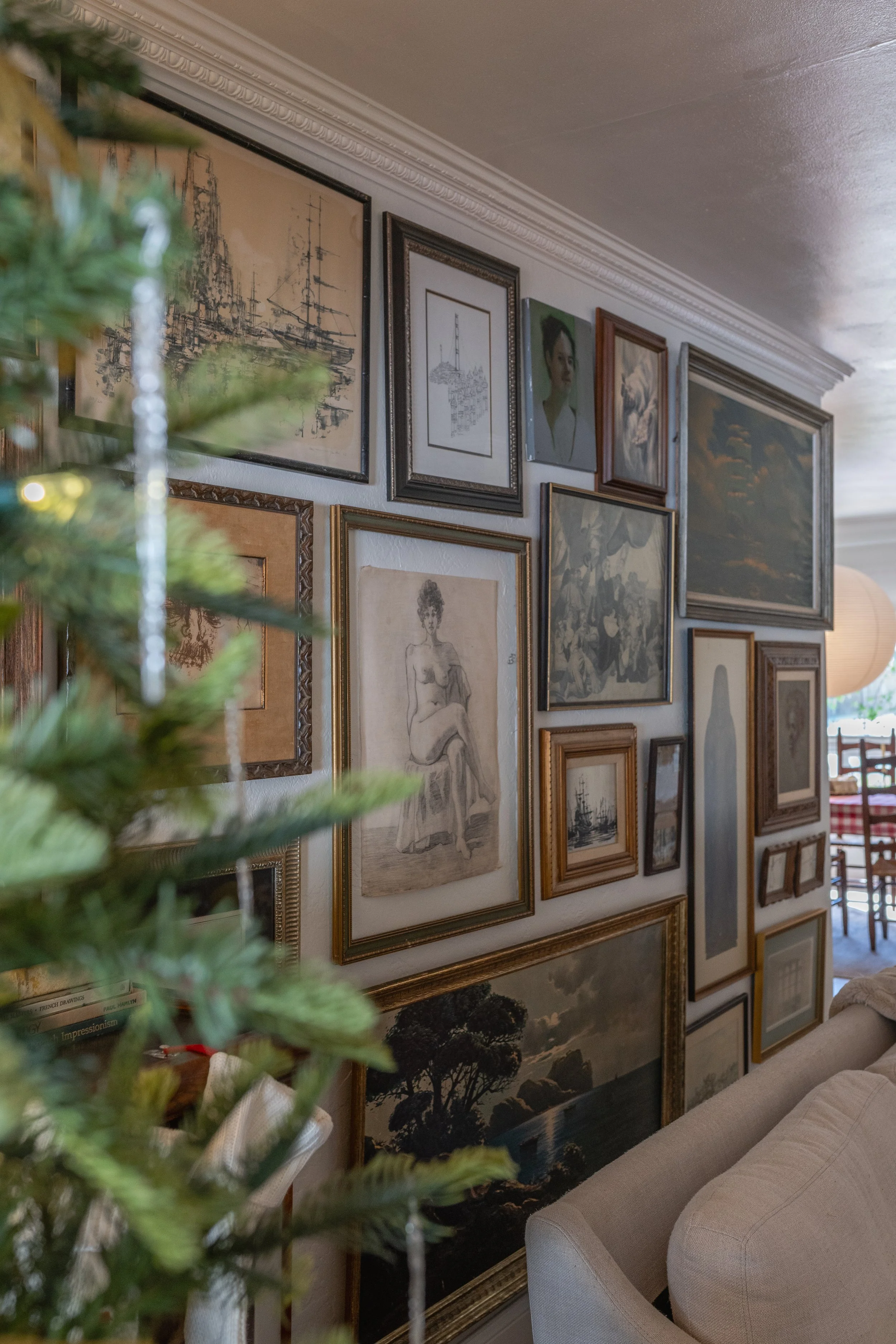 A wall decorated with various framed artwork and photographs, with part of a Christmas tree and a beige sofa visible in a cozy living room.