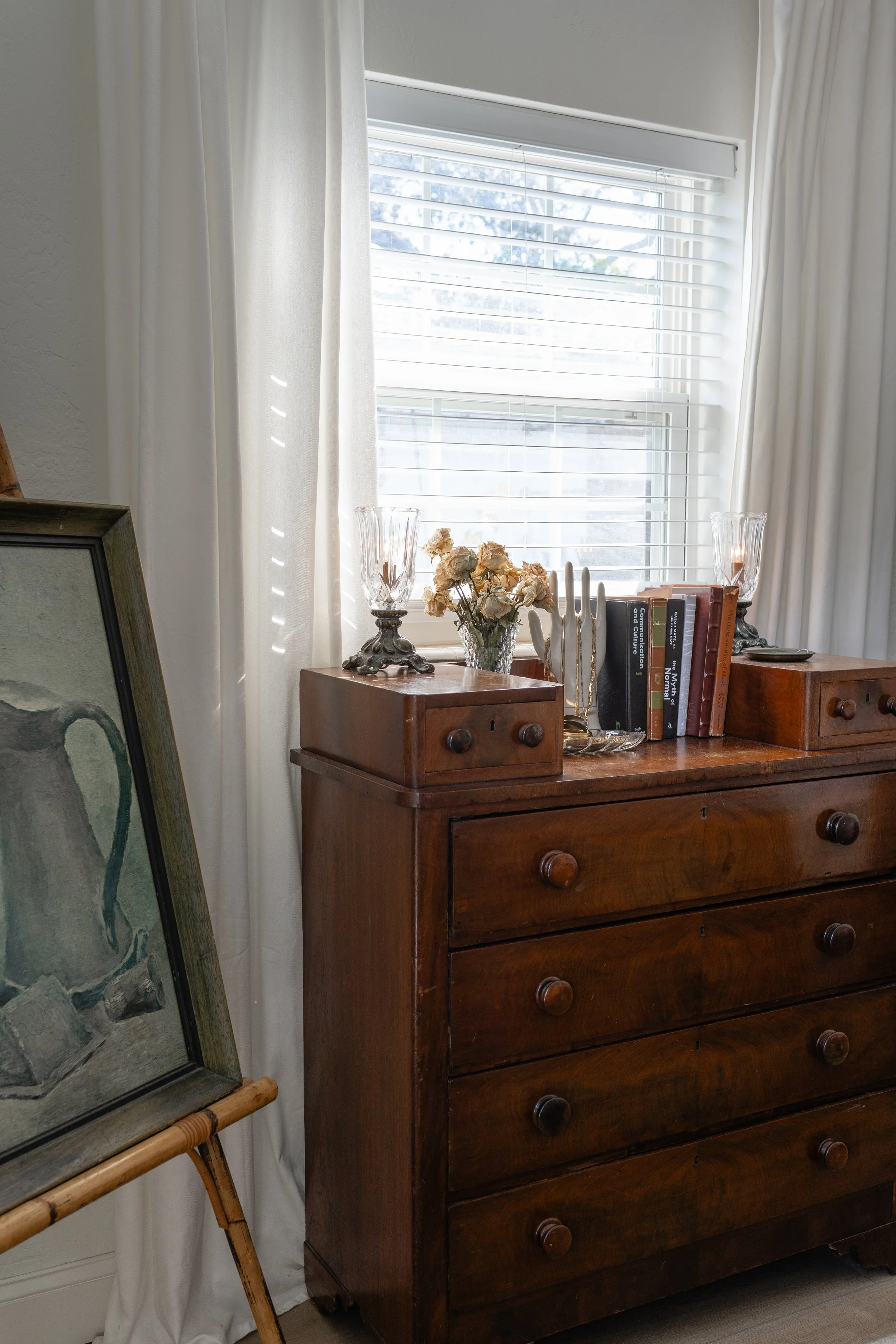 A wooden dresser with rounded knobs, decorated with a glass vase of dried flowers, two candle holders, a few books, and decorative gold-colored objects, placed in front of a window with white blinds and curtains. Part of an easel with a framed artwor