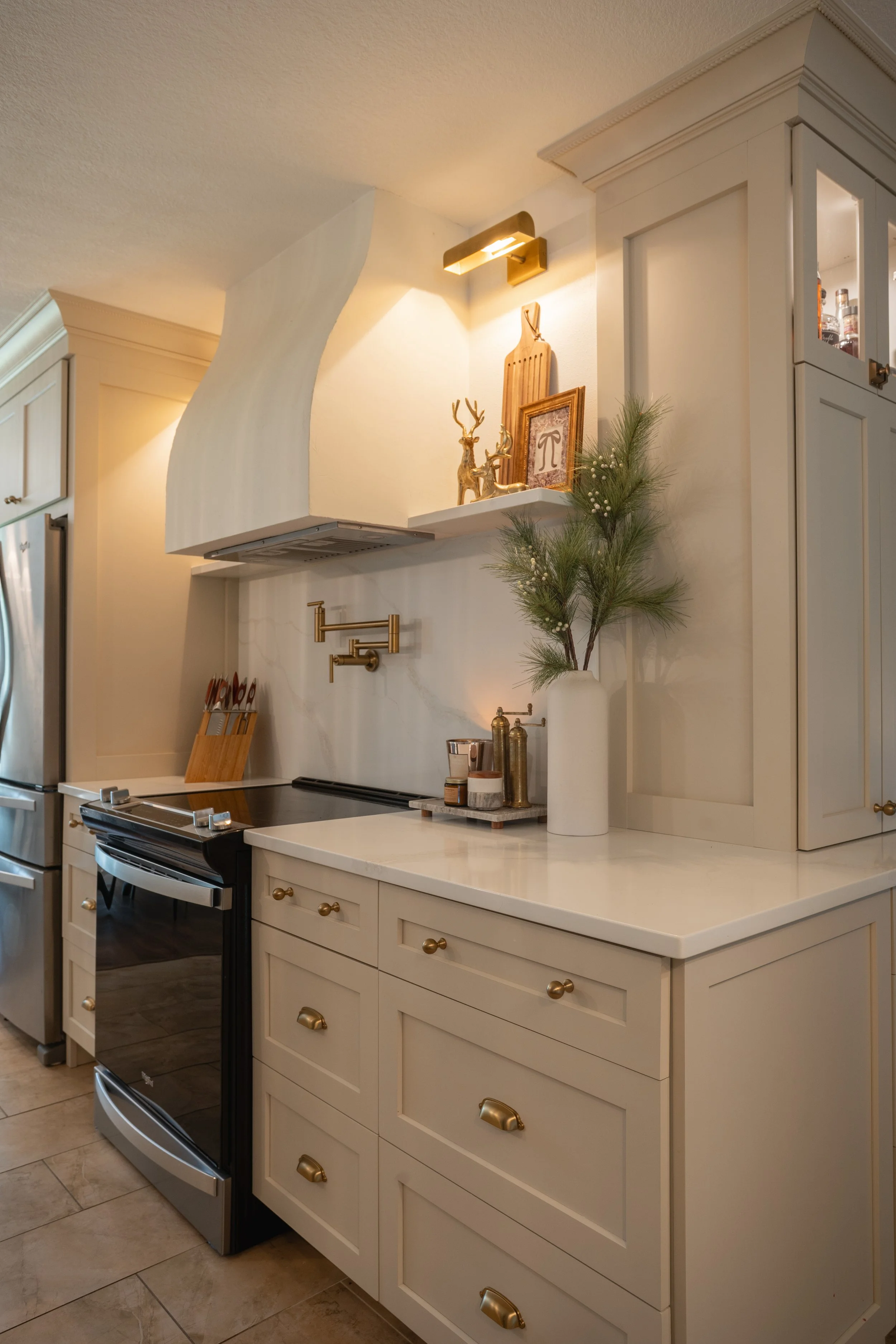 A cozy kitchen corner decorated with green pine branches in a white vase, gold hardware, a cutting board, and festive ornaments on a shelf near a stove and refrigerator.