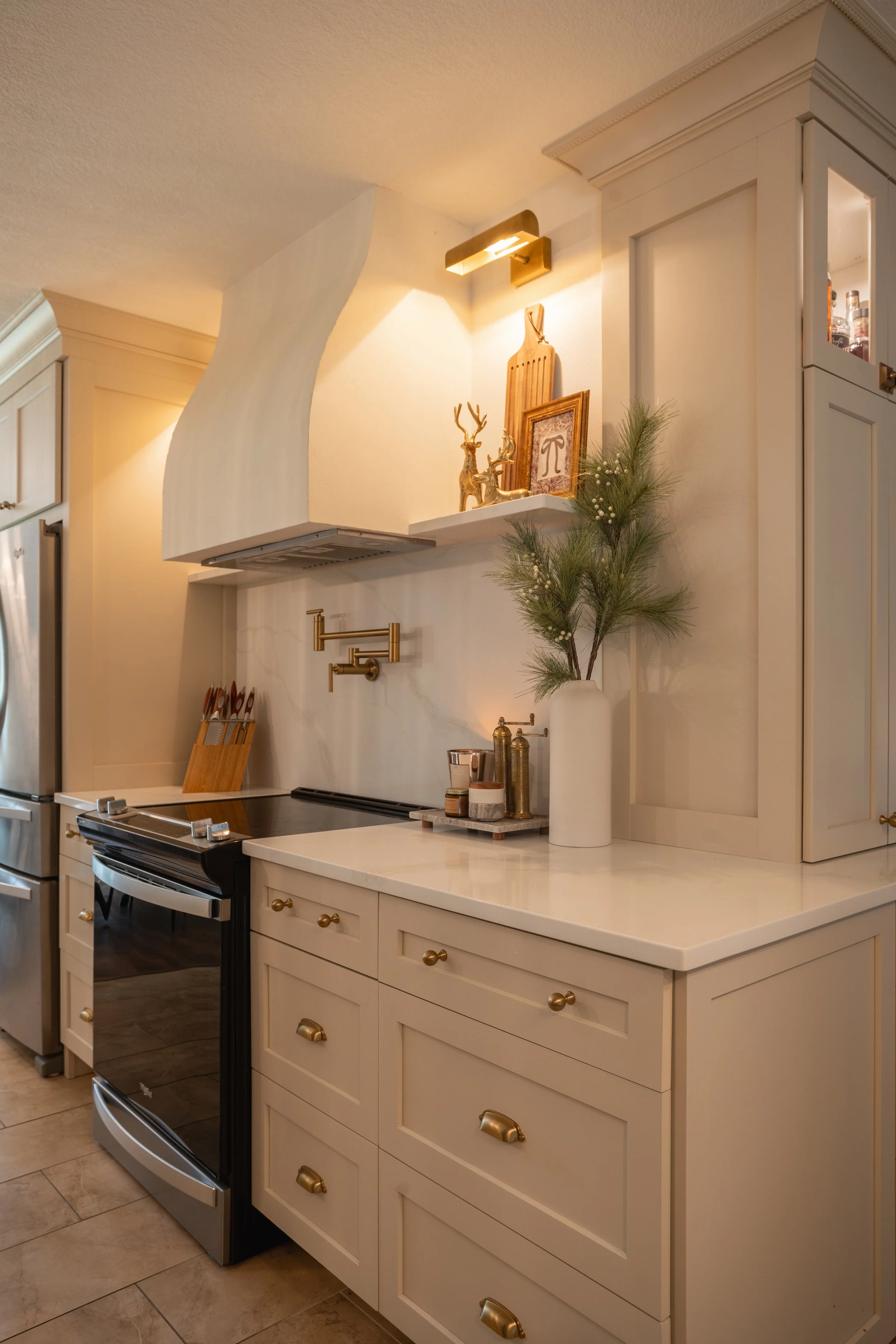 Kitchen with cream-colored cabinets, a white countertop, and decorative holiday accents including a vase with pine branches, gold figurines, and framed art. A black oven and stainless steel refrigerator are visible.