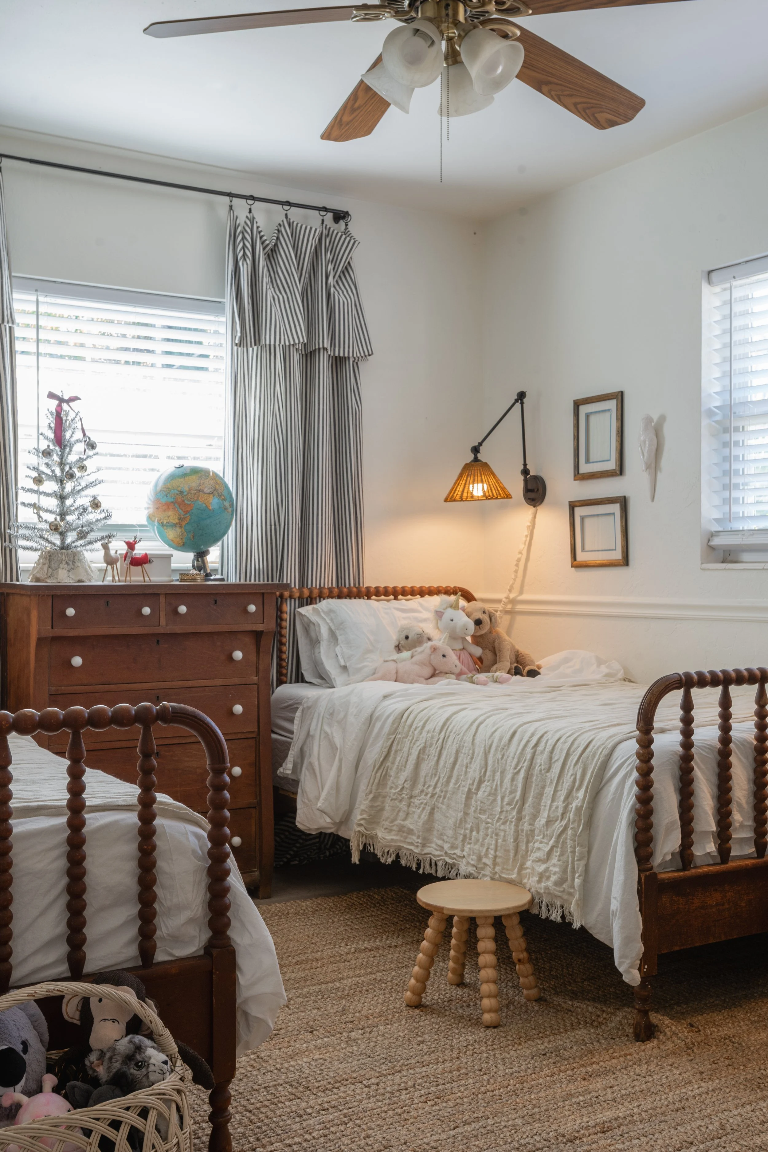 Bedroom with two wooden beds, stuffed animals on one bed, dresser with a globe and holiday decorations, small wooden stool, striped curtains, ceiling fan, and framed wall art.