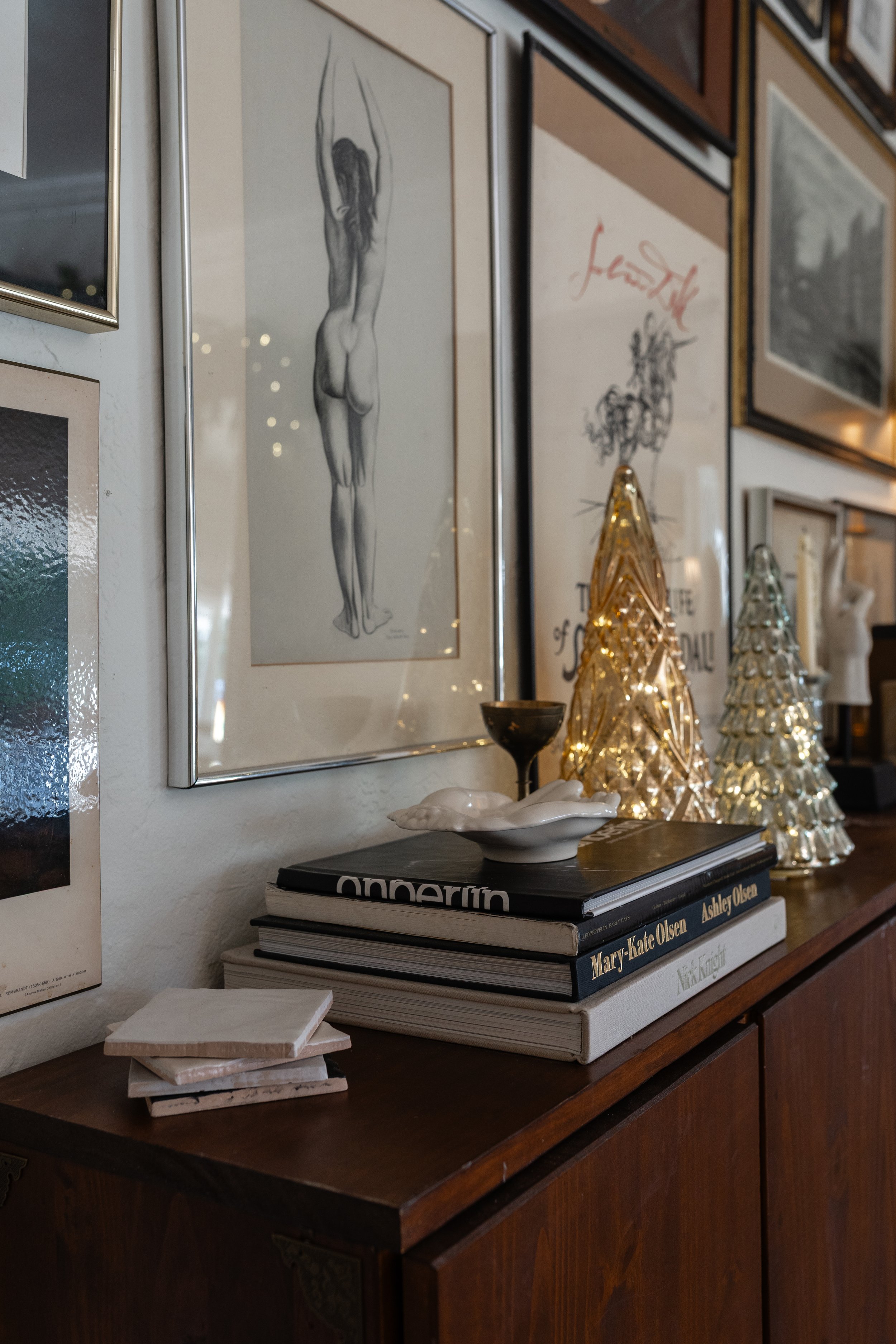 Decorative display on a wooden sideboard with framed art, books, and Christmas trees with string lights.