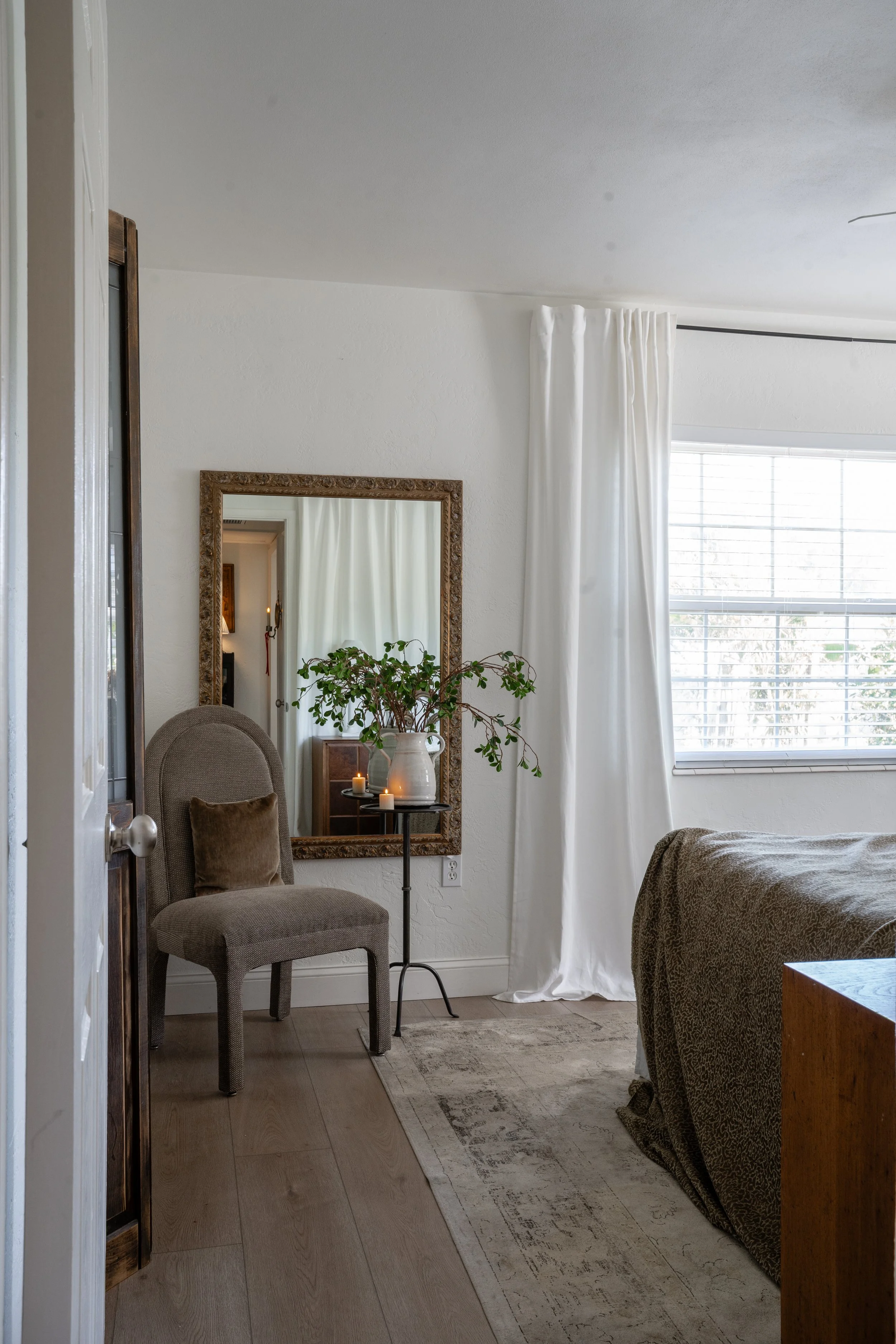 A cozy bedroom corner with a beige upholstered chair, a mirror with an ornate gold frame reflecting part of the room, a small black table with a large white vase holding green leafy branches, and three small candles. White curtains frame a window with blinds, and part of a bed with a patterned blanket is visible.