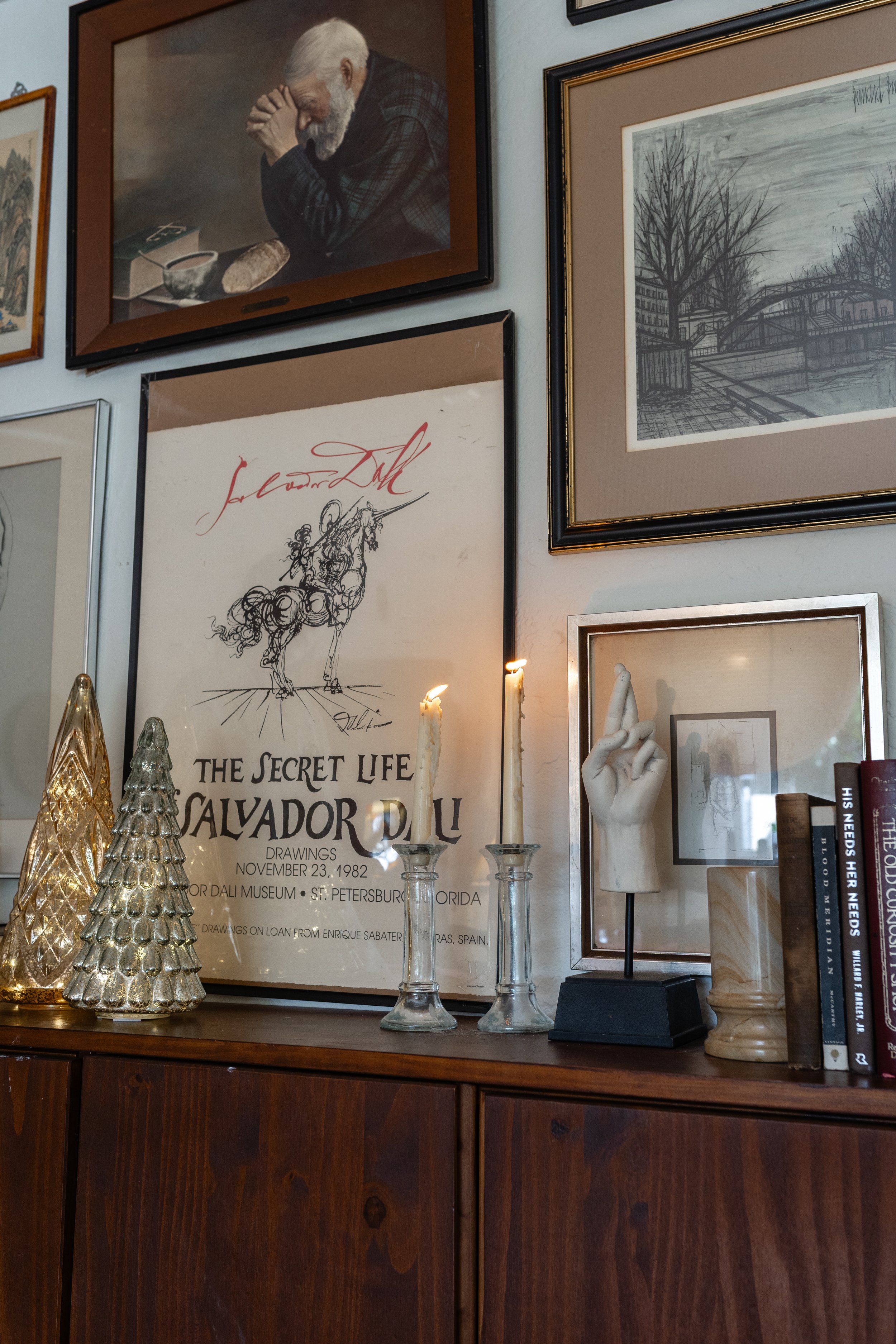 A gallery wall with various framed artworks, including a portrait of an elderly man with a beard praying, a landscape drawing, a poster for Salvador Dali artworks, and an abstract sculpture of a hand, with decorative trees and books on a wooden surfa