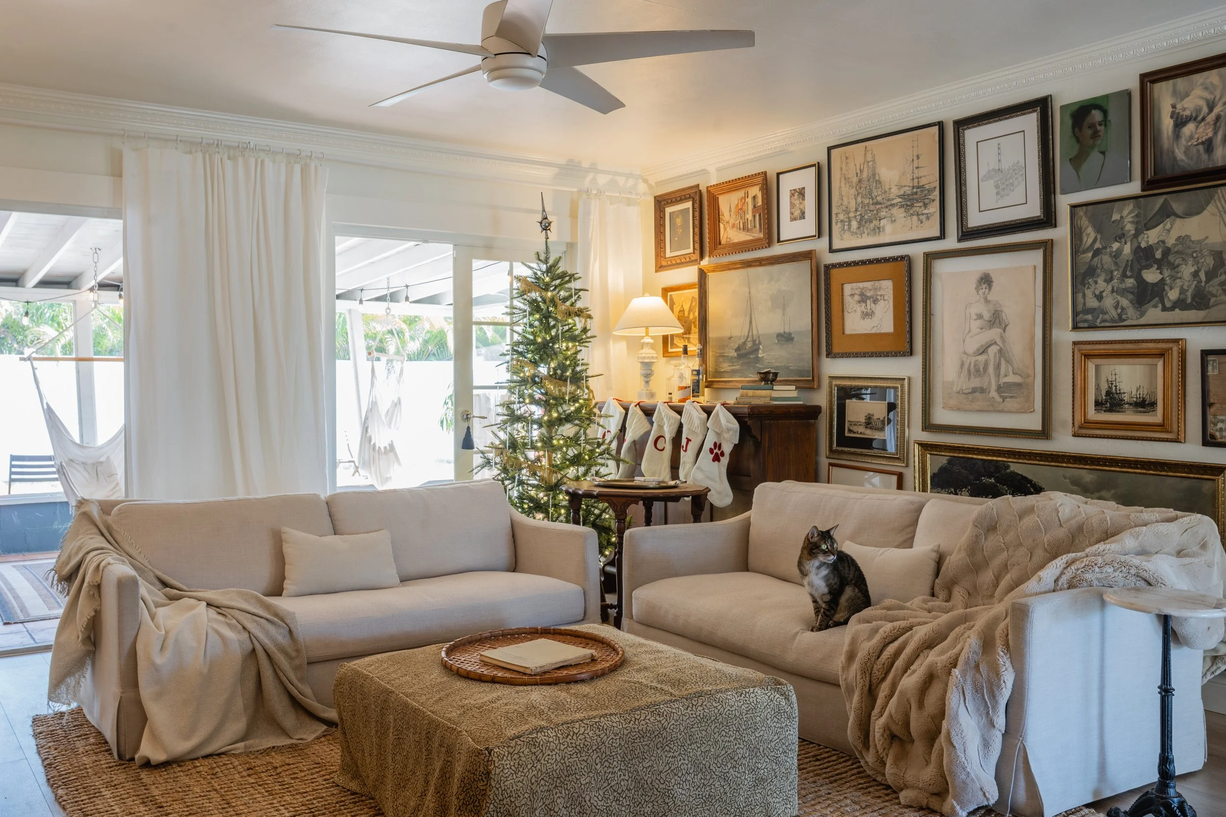 Living room decorated for Christmas with two beige sofas, a small Christmas tree, a gallery wall of framed artwork, and a cat sitting on one sofa, with sheer curtains and glass sliding door leading to a patio with a hammock.