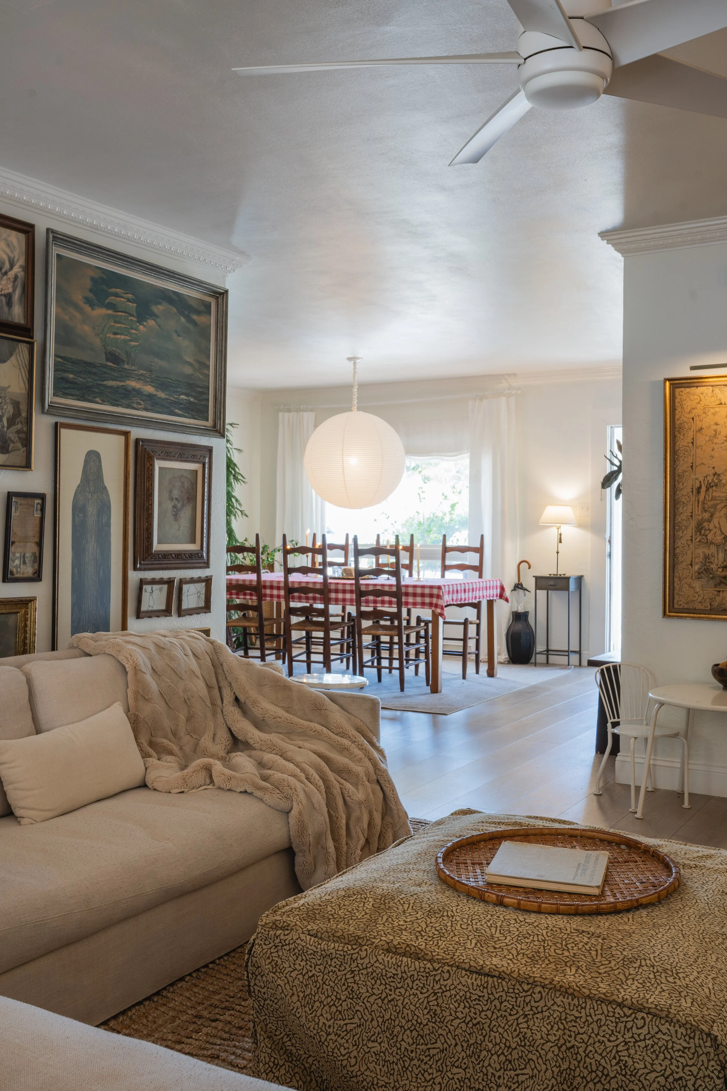 Living room with beige sofa, fur blanket, coffee table with a book, gallery wall, and dining area with a red checkered tablecloth, wooden chairs, hanging lantern, and window with curtains.