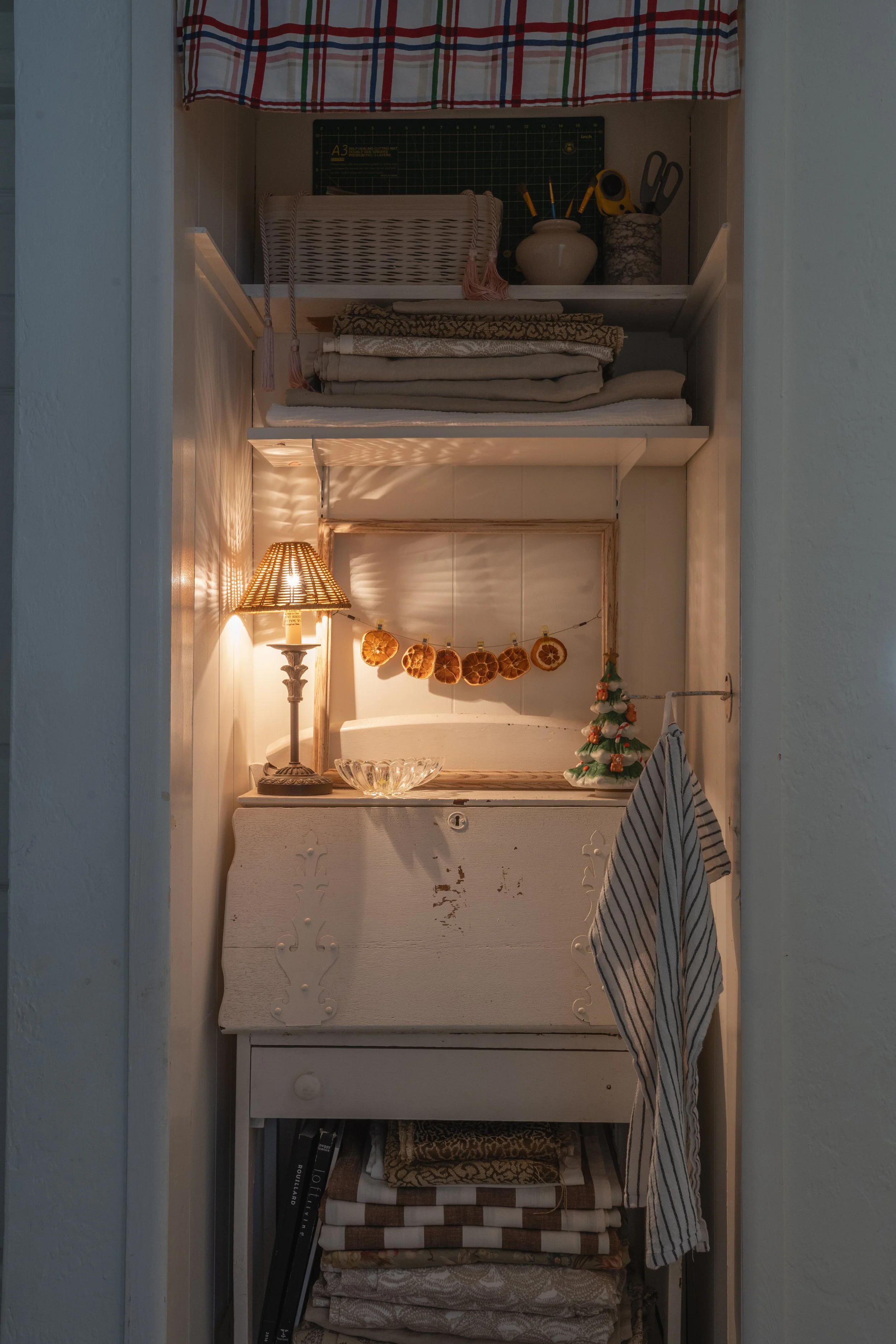 Tall, narrow cabinet filled with folded linens, a small table with a lamp, dried orange slices hanging on a string, a small decorative tree, and a glass bowl.