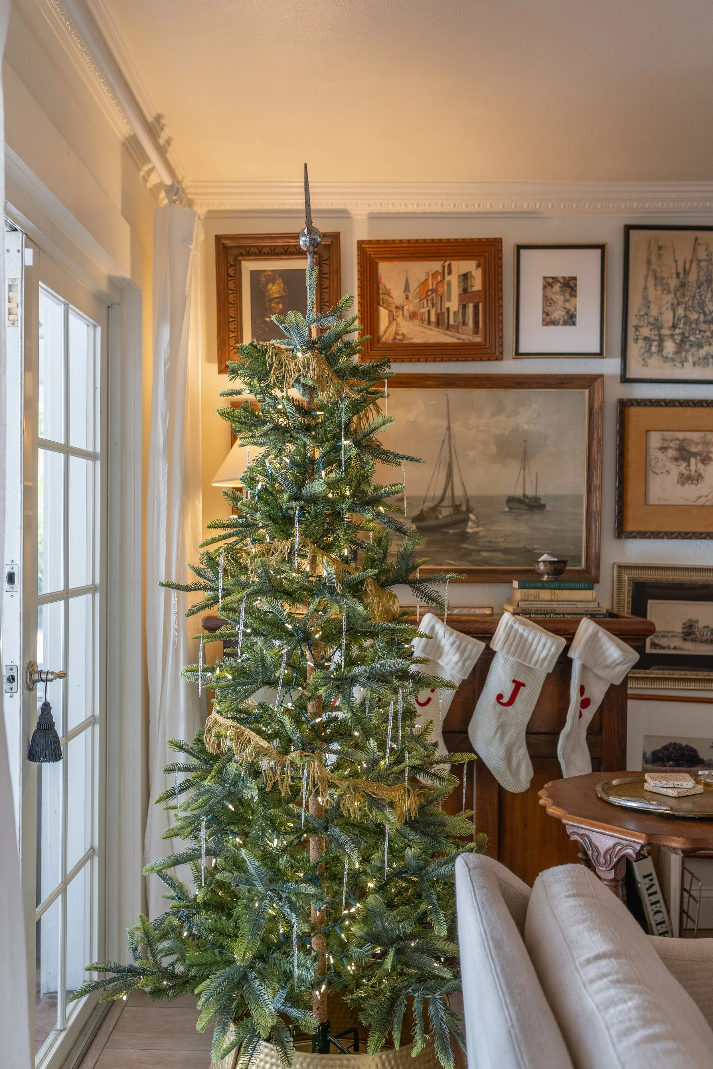 A decorated Christmas tree in a living room with framed artwork on the wall, a table with books, and stockings hanging.