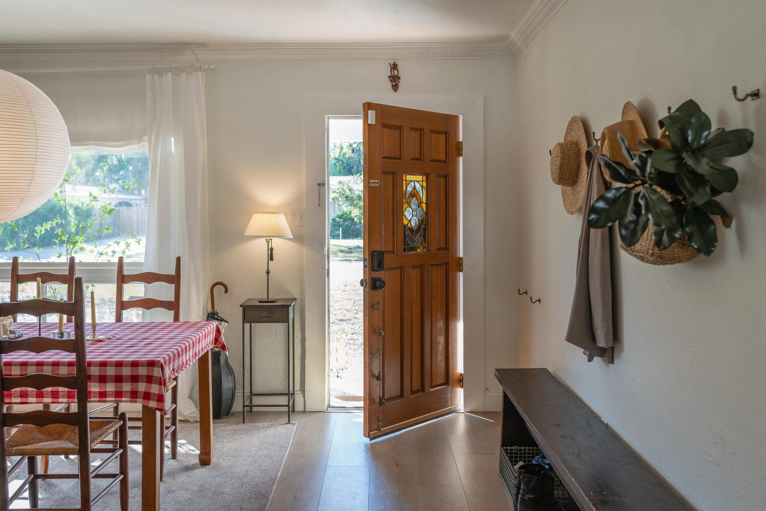 Interior view of a home entryway featuring a wooden front door with stained glass window, open to outside. To the left, a dining area with a red and white checkered tablecloth, wooden chairs, also a lamp on a small side table. To the right, a wall wi