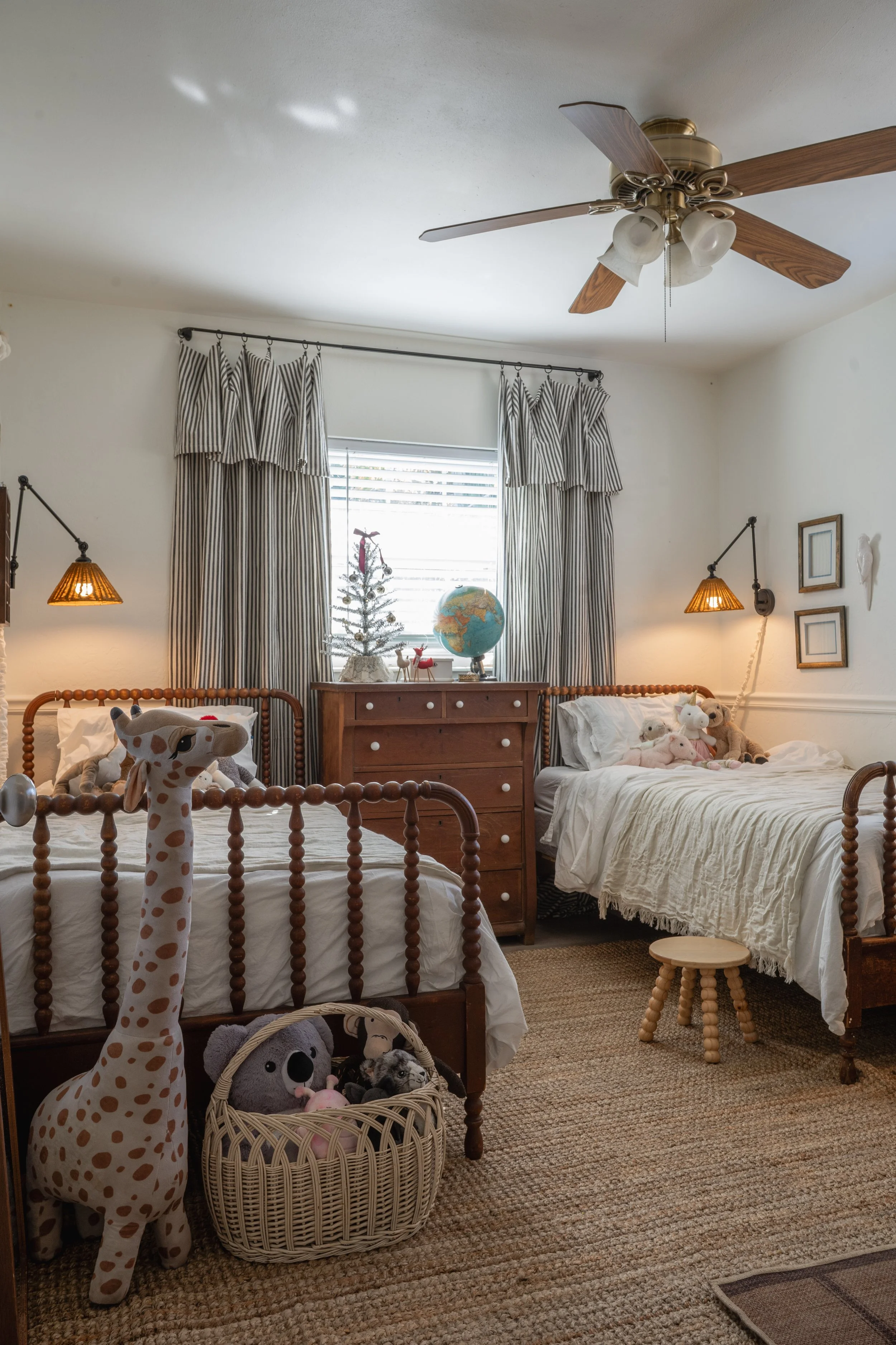 A cozy children's bedroom with two twin beds, stuffed animals, a small wooden stool, and a dresser with a globe and decorative items. The room features a ceiling fan, striped curtains, and framed art on the wall.