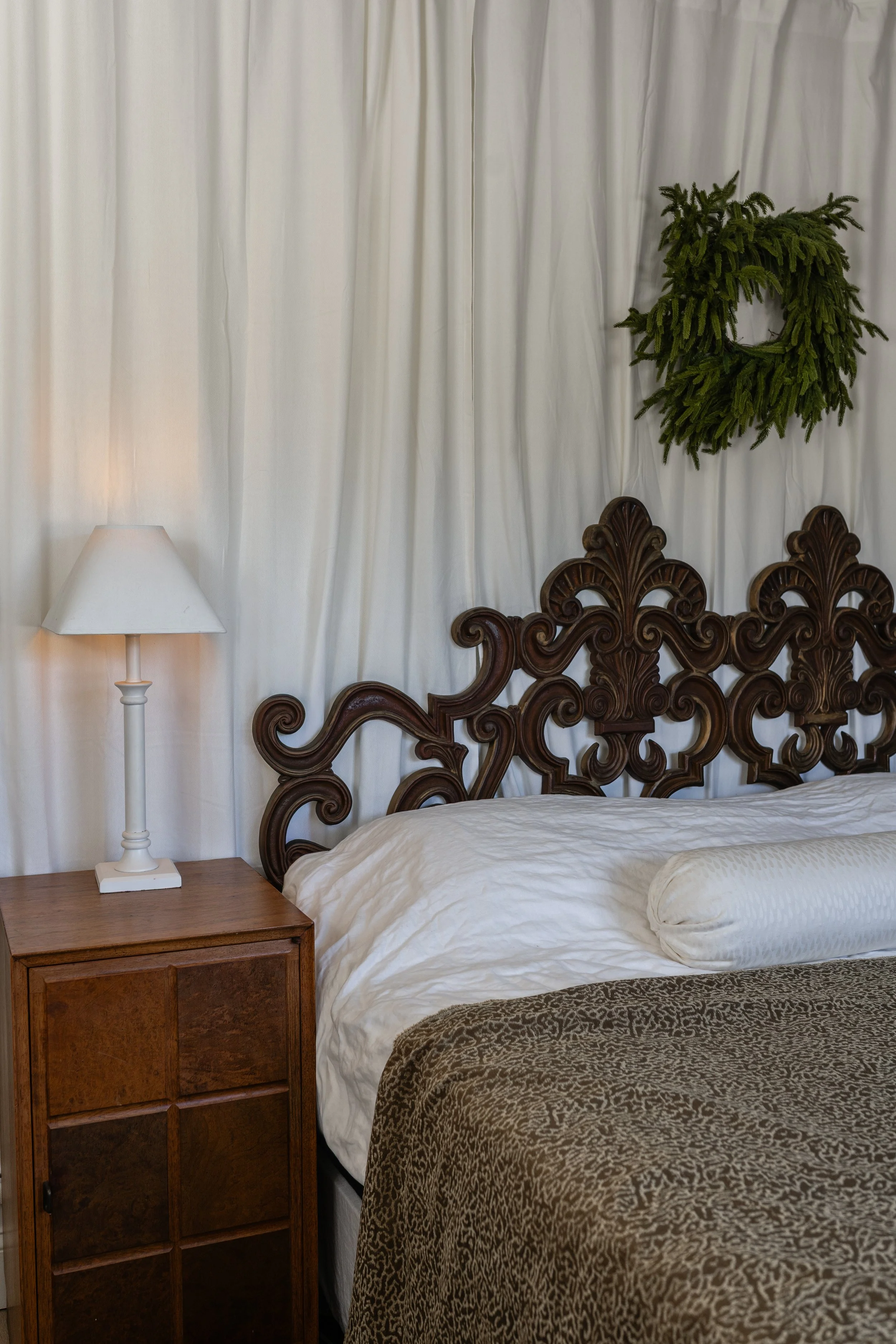 Close-up of a decorated bedroom with a white lamp on a wooden bedside table, a carved wooden headboard, white curtains, and a green wreath on the wall.