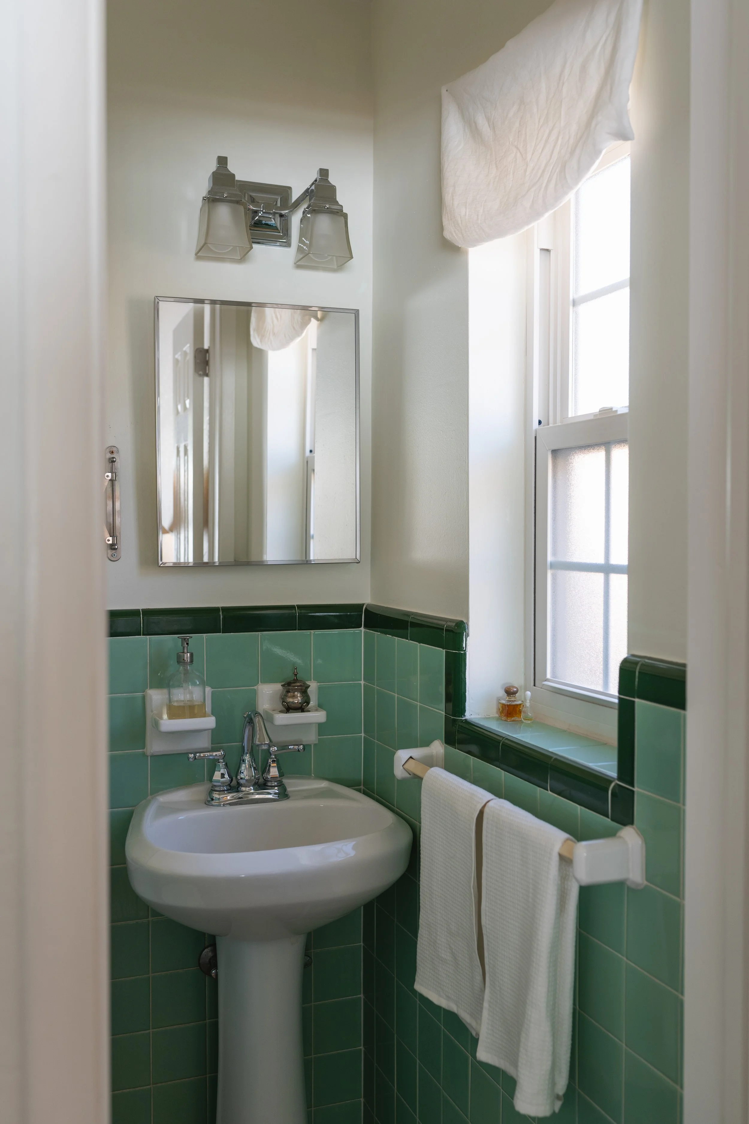 A vintage bathroom with green tiled walls, a pedestal sink, a small mirror, a towel rack with a white towel, a window with a cream curtain, and an overhead light fixture.