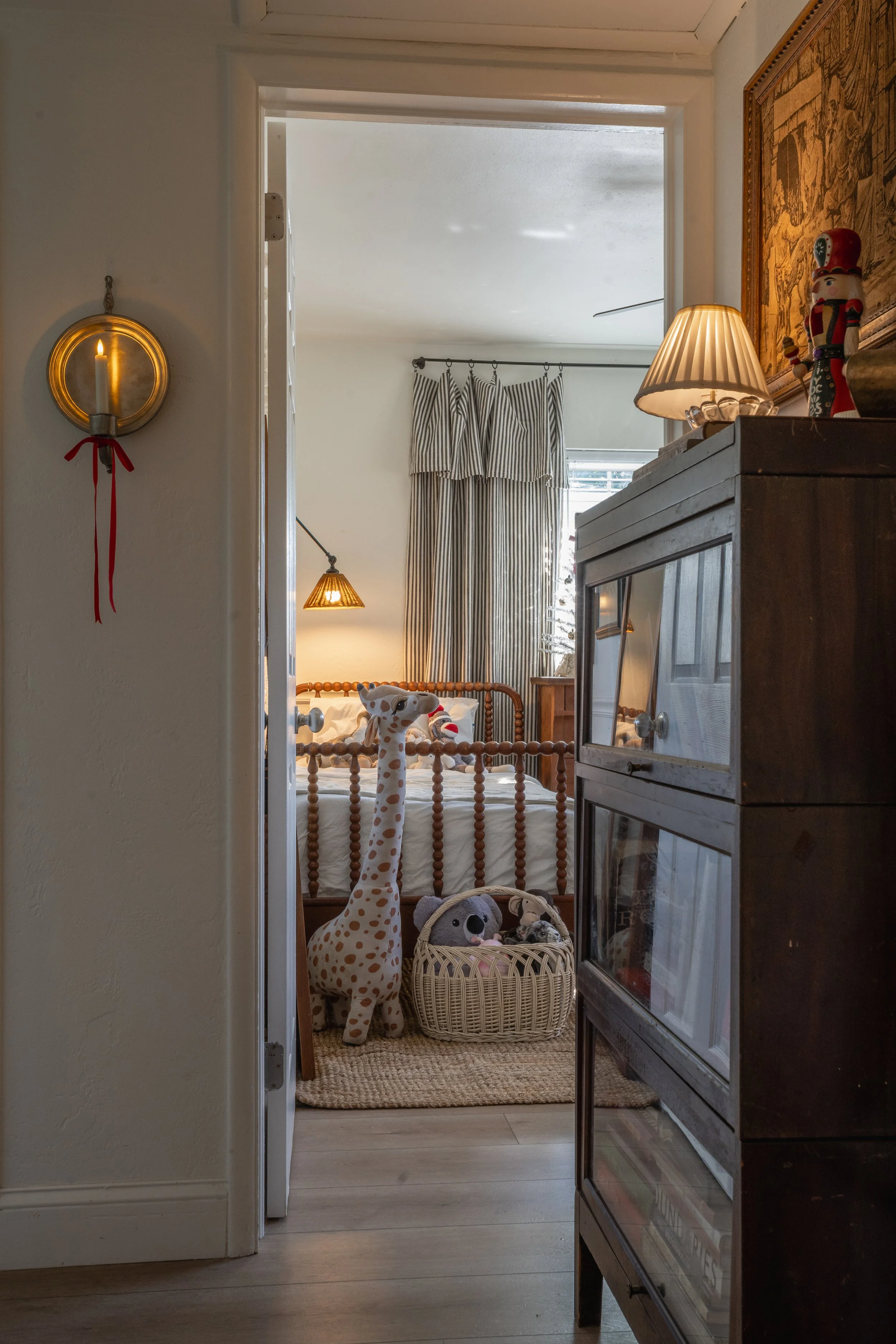 View into a child's bedroom with a wooden bed, striped curtains, and various plush toys including a giraffe, koala, and other stuffed animals in a basket.