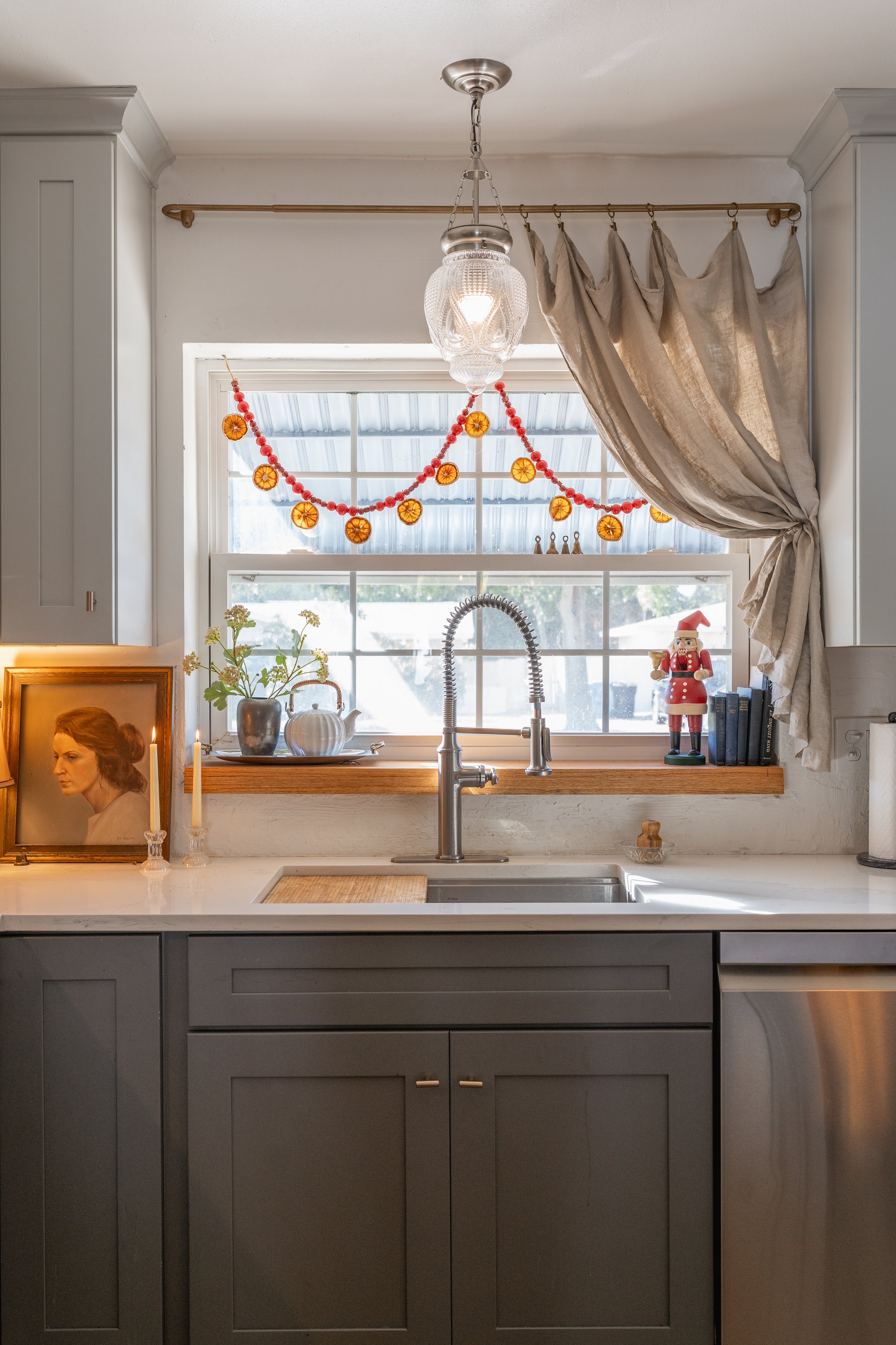 A kitchen window decorated with holiday garland and ornaments, featuring a sink, faucet, and various decorative items on the windowsill, including a framed portrait, candles, a teapot, vases, and a nutcracker figurine.
