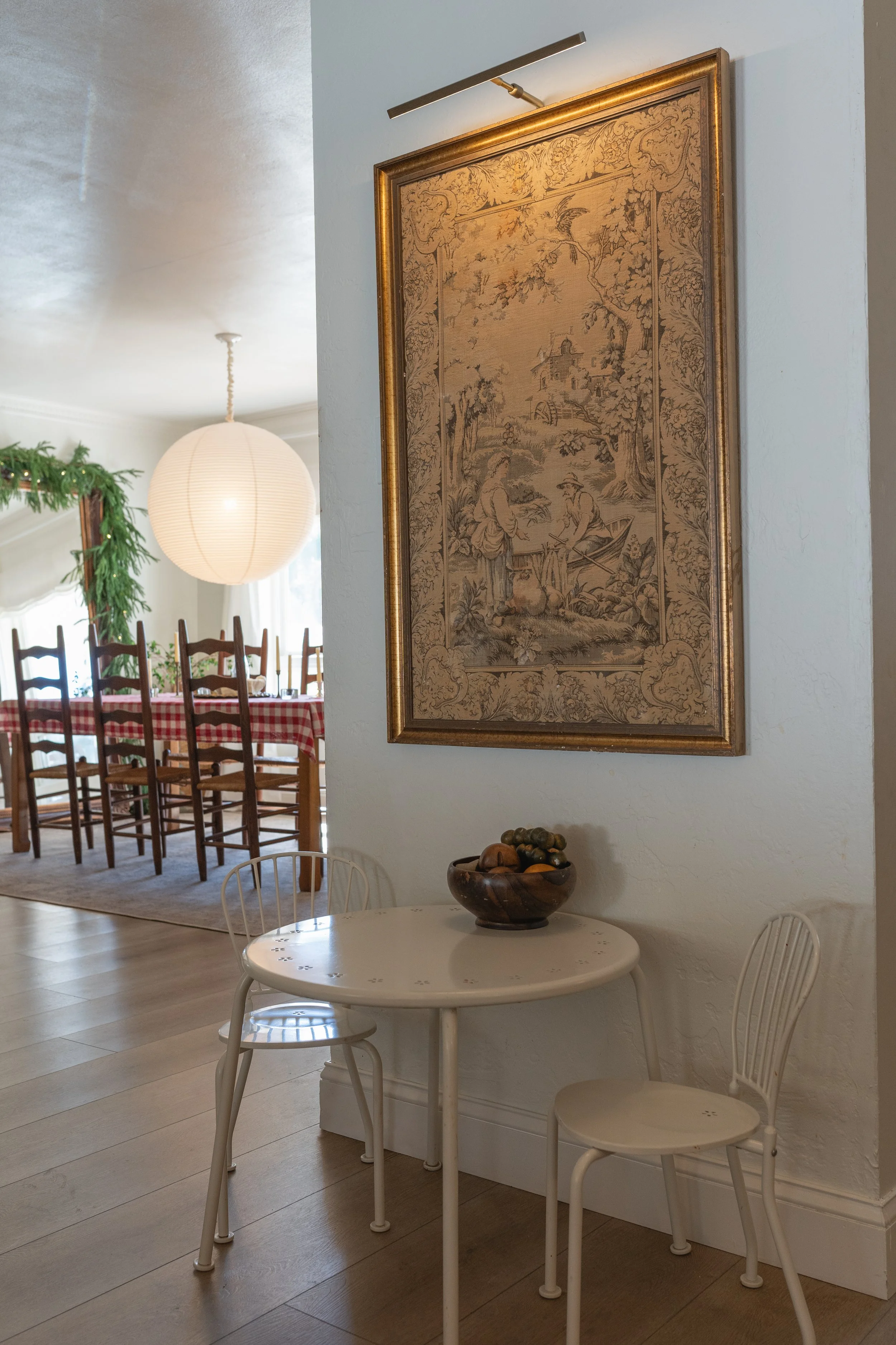 A small white round table with two matching white chairs, a wooden bowl with fruit, an elaborate framed tapestry hanging on the wall, and a dining area with a long table covered with a red plaid tablecloth in the background.